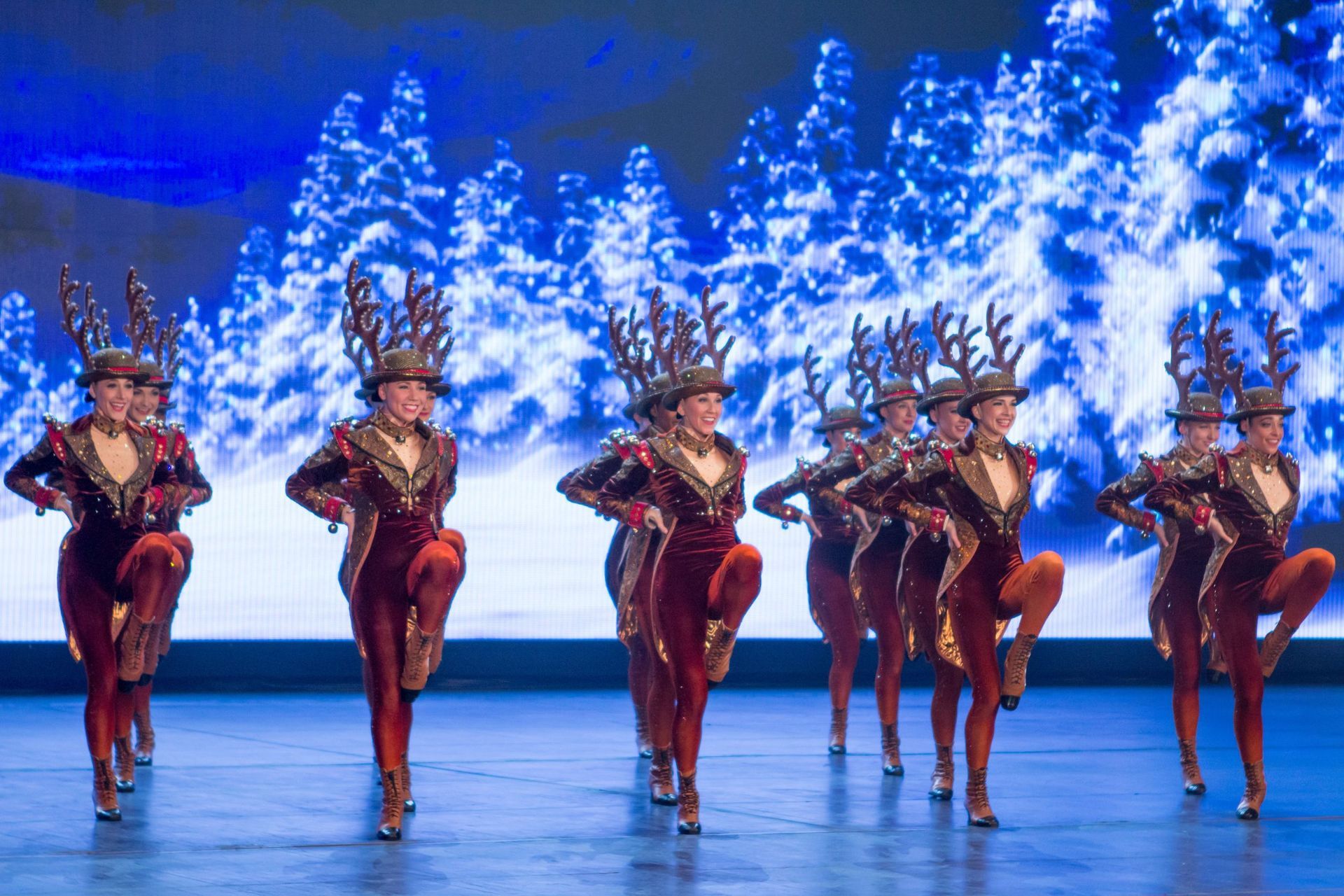 Dancers in reindeer costumes perform on stage in front of a winter forest backdrop.