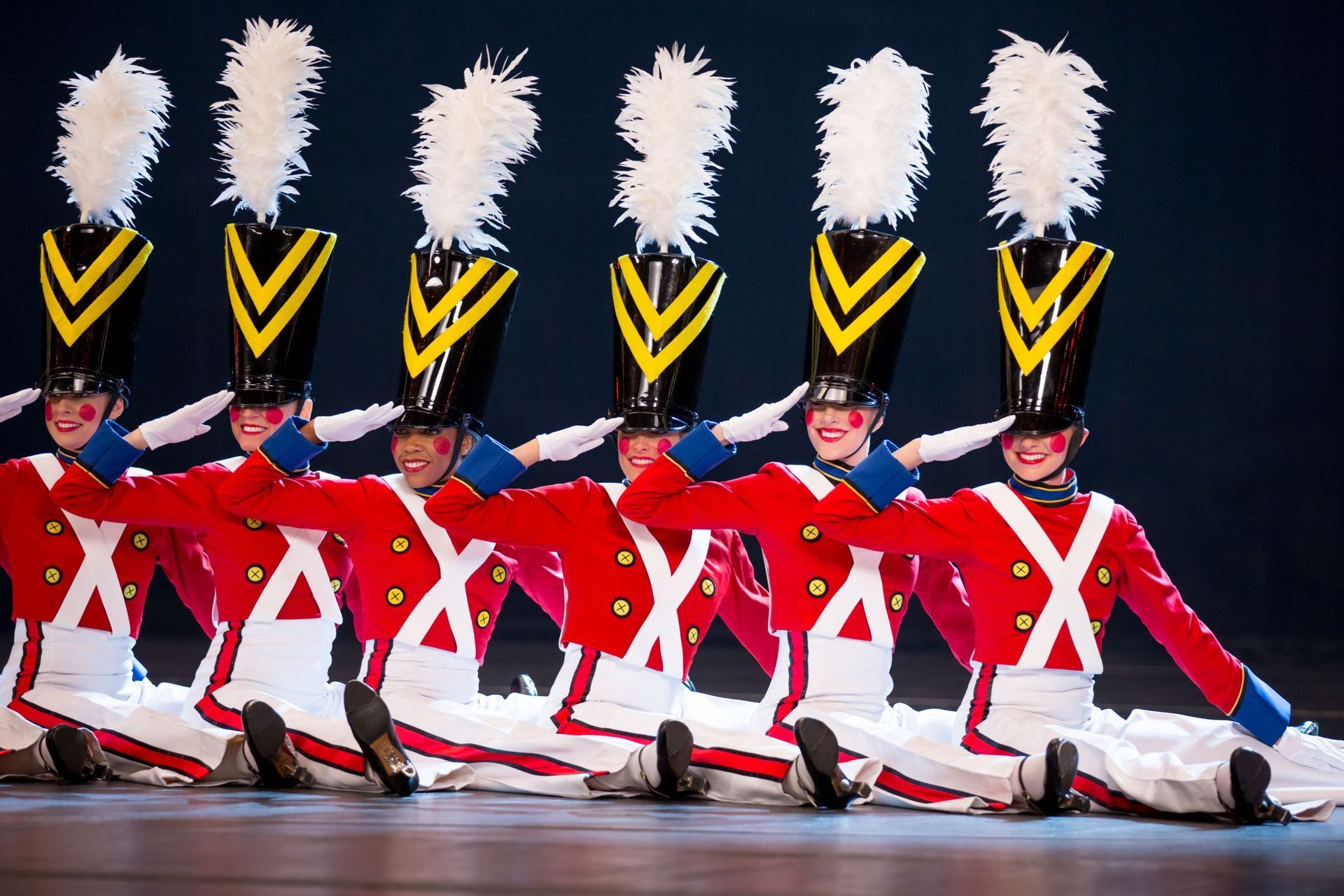 Toy soldier dancers in red and white costumes, saluting, with tall black hats and feathered plumes.