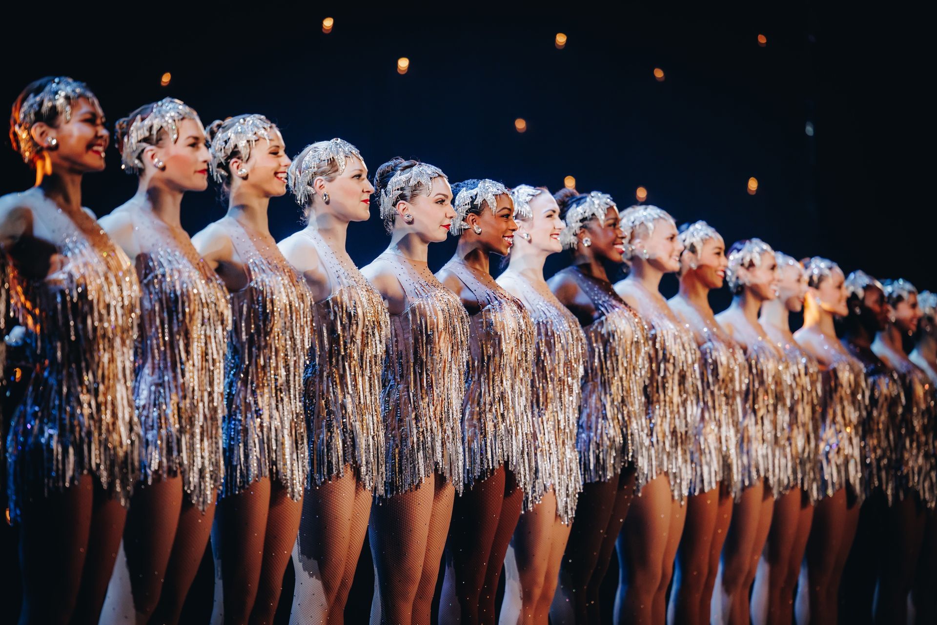 Dancers in glittering gold costumes, lined up on stage, looking toward the audience.