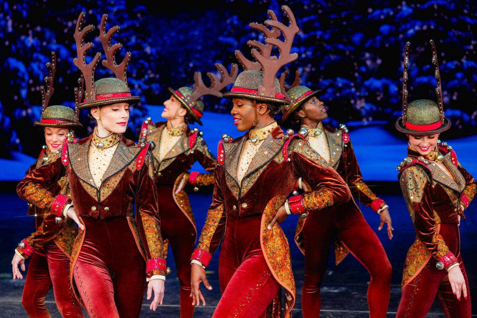 Dancers in velvet costumes with antlers perform onstage; red, gold, and green against a blue backdrop.