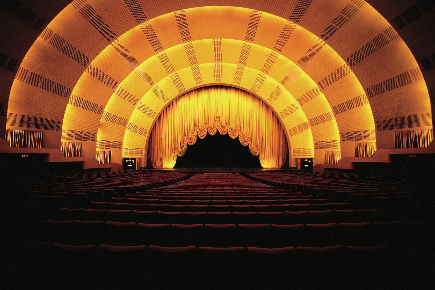 Interior view of the Radio City Music Hall stage, lit in gold with rows of red seats in the foreground.