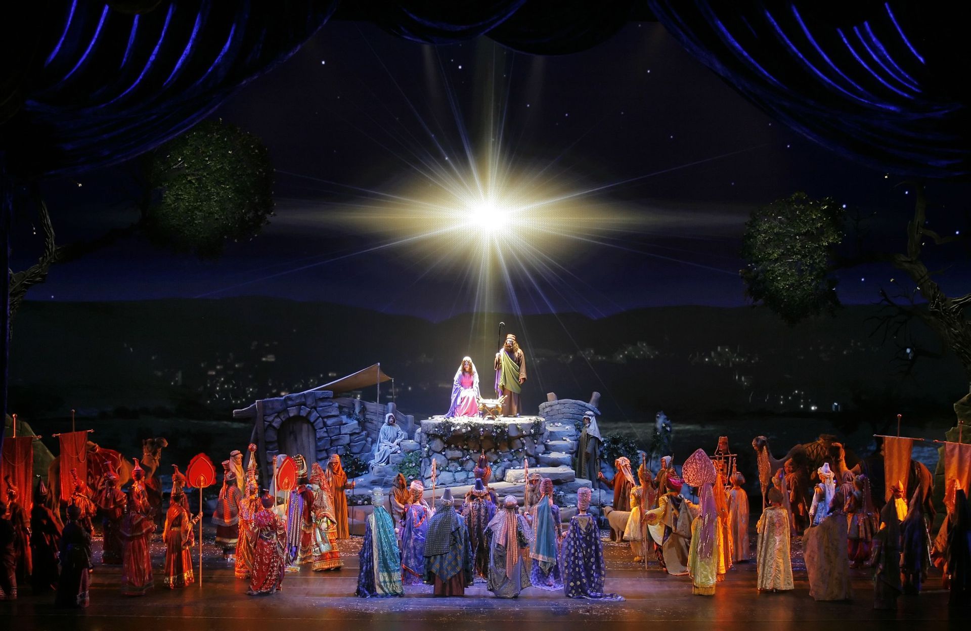 Nativity scene on stage with many costumed figures, bright light above a manger.
