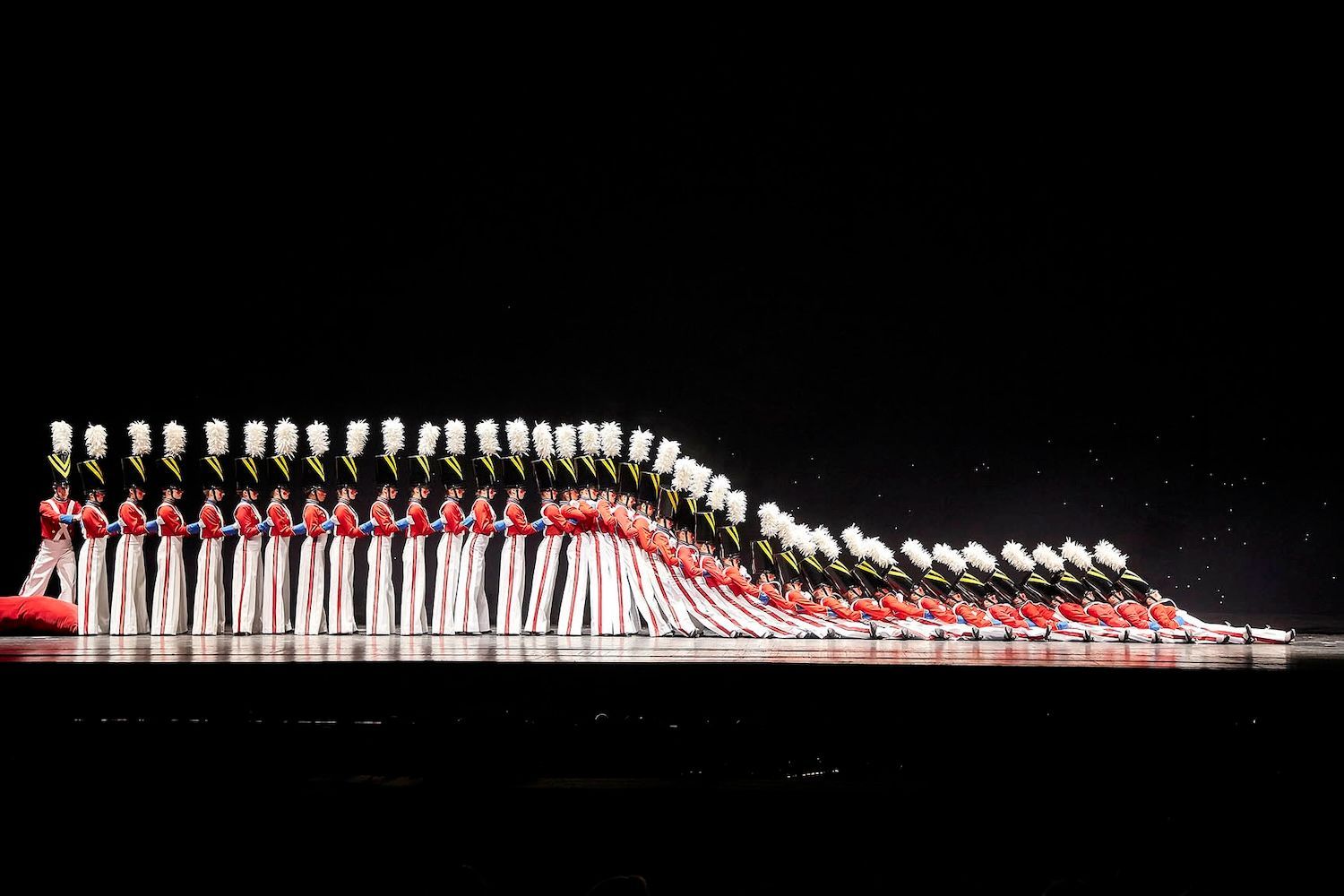 A marching band performs on a stage, their white and red uniforms and white hats creating a wave-like formation.