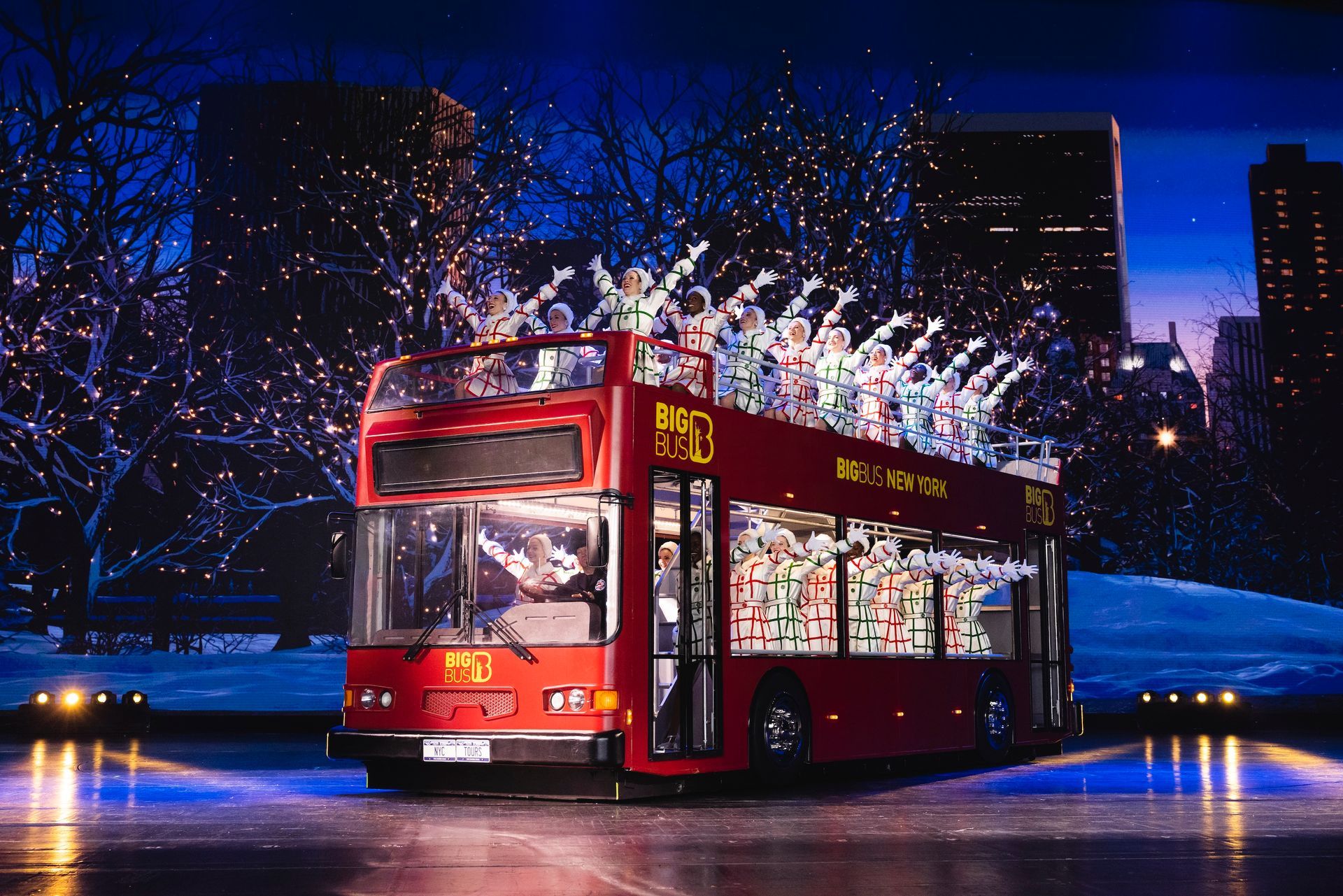 A red double-decker bus on stage with a nighttime cityscape backdrop and lights.