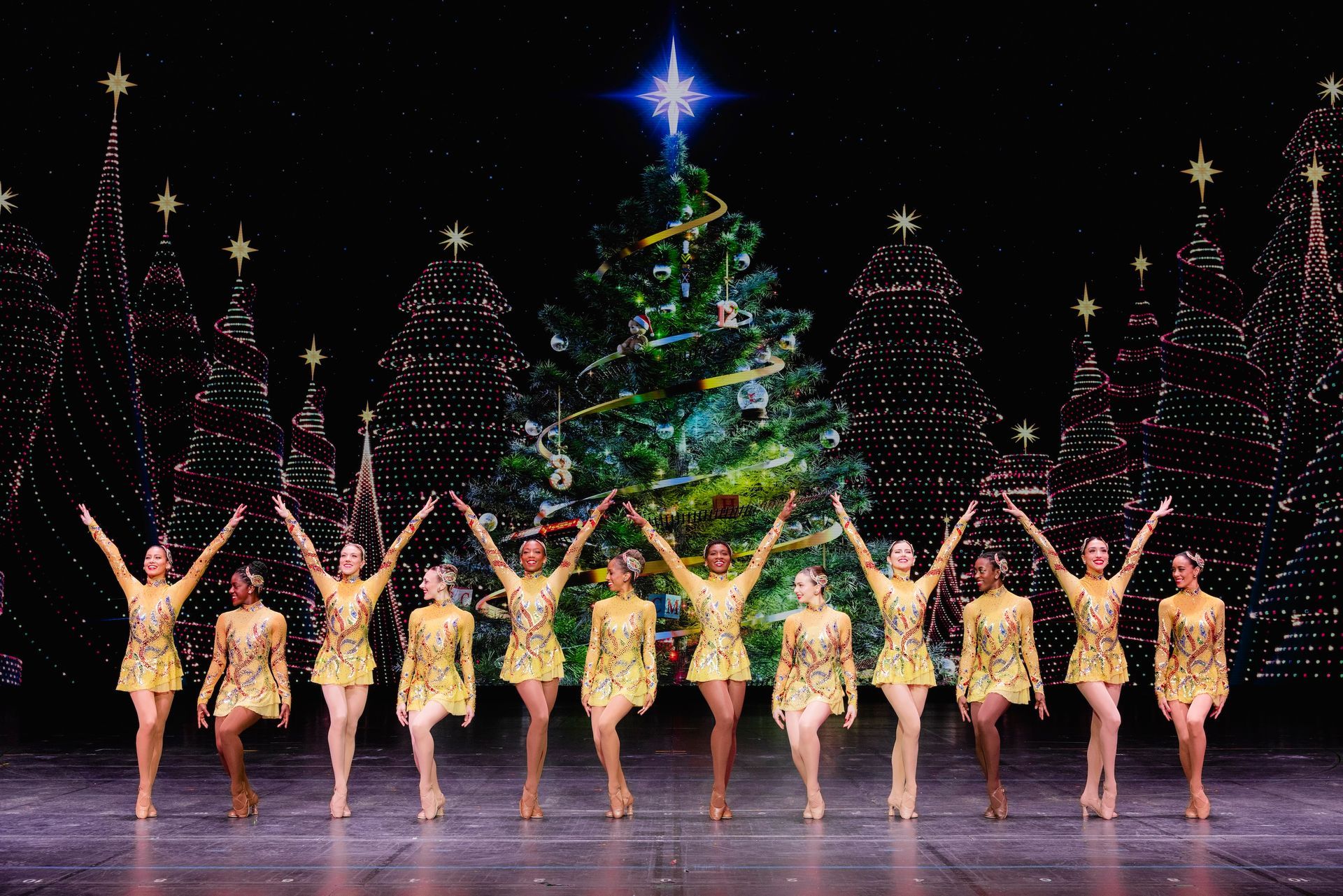 Dancers in gold costumes with arms raised, Christmas tree backdrop.