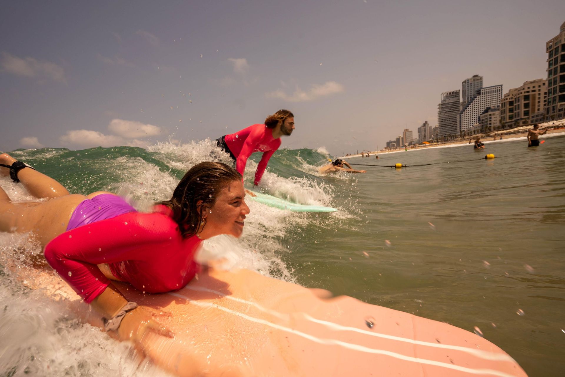 Woman and another surfer paddling on surfboards in ocean, sunny day at beach with buildings in background.