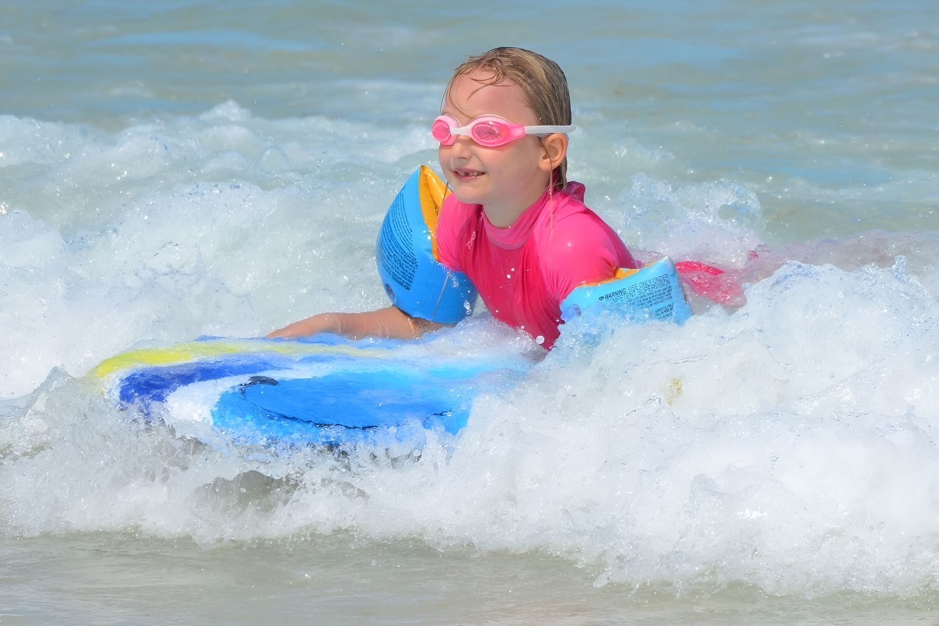 Young girl, wearing pink goggles, swimsuit, and arm floats, surfs a wave on a blue bodyboard.