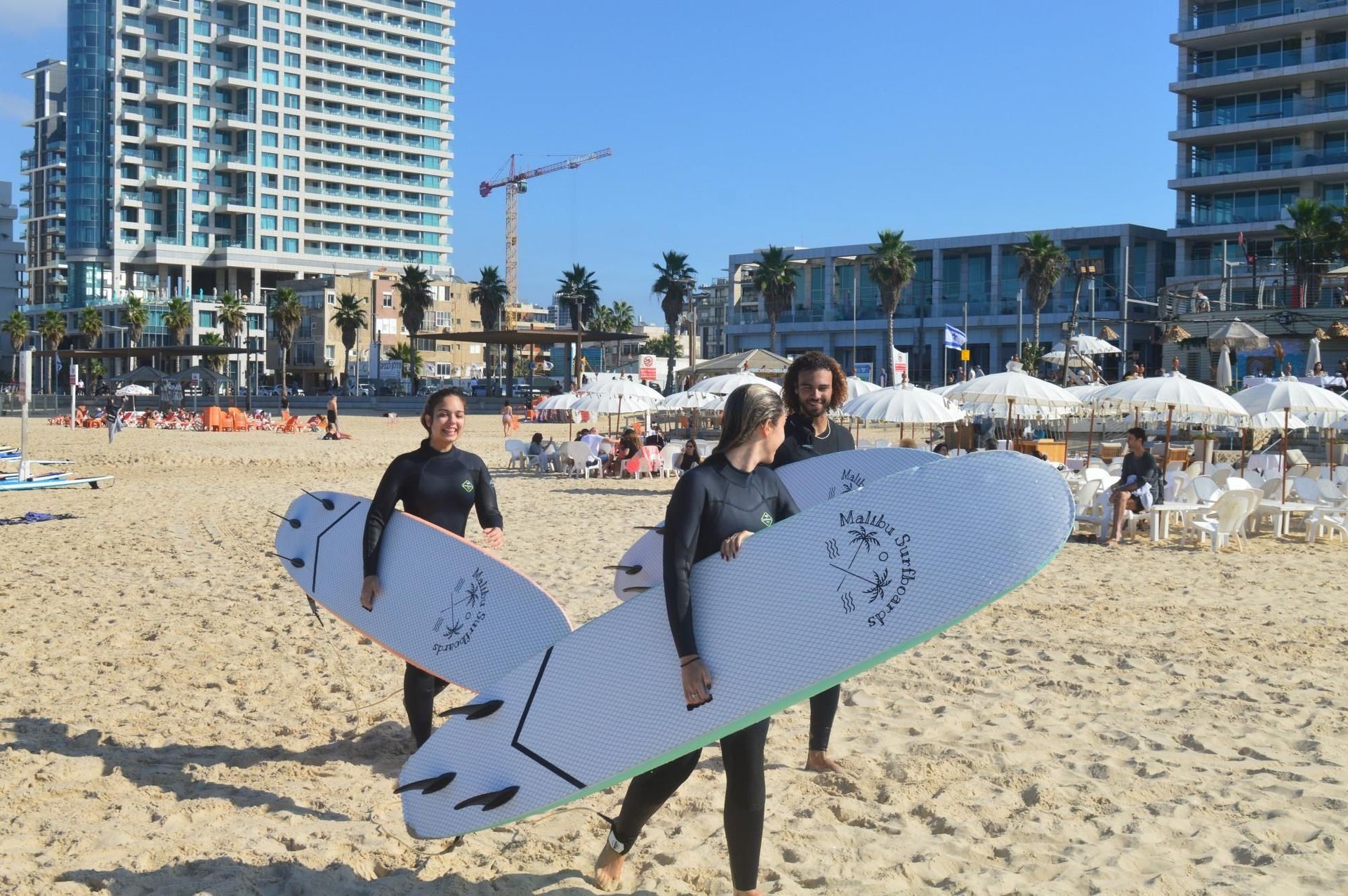 Three people in wetsuits carry surfboards on a sunny beach. City buildings and umbrellas are in the background.