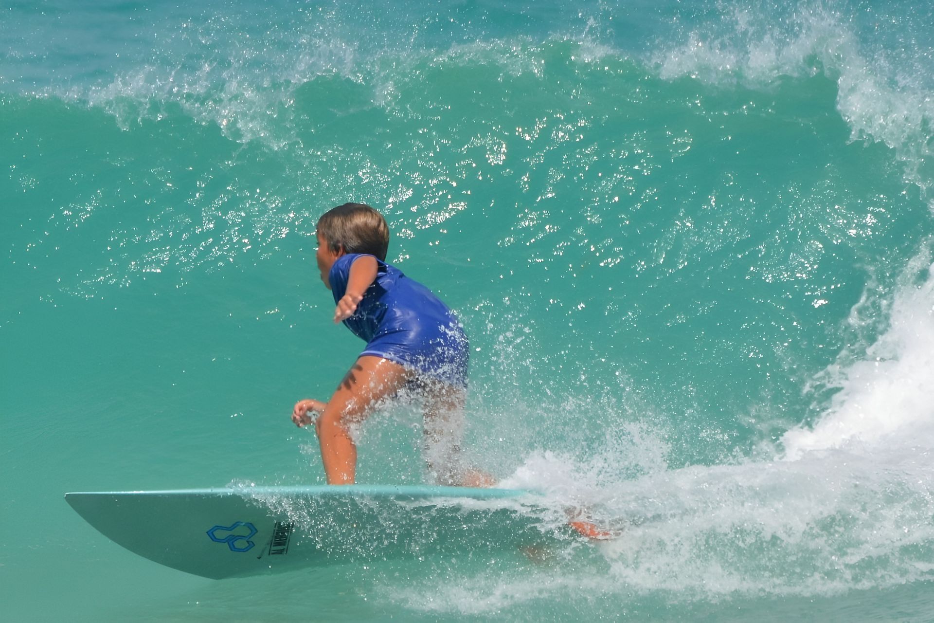 Young surfer in blue suit riding a turquoise wave, ocean setting.