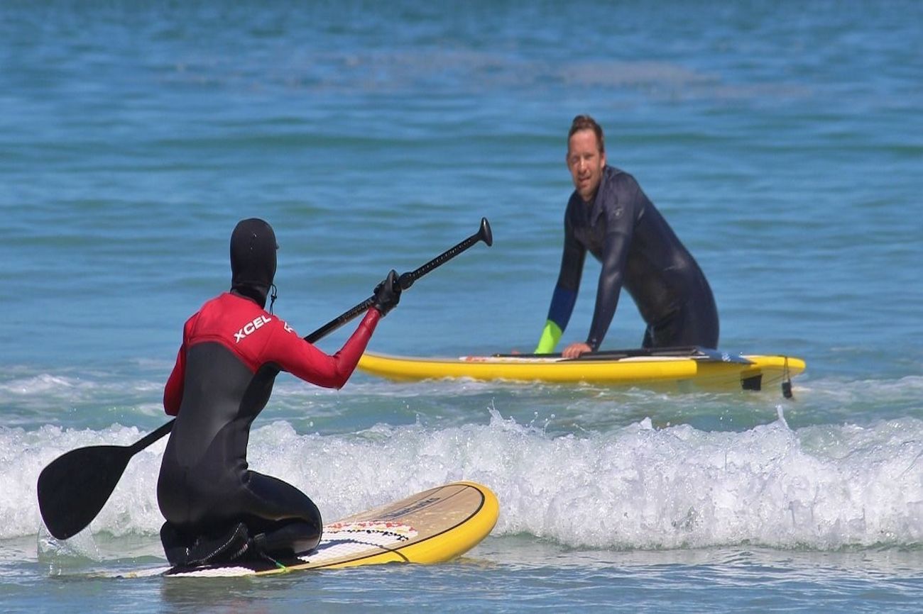 Two people on paddleboards in the ocean. One kneels with a paddle, the other stands in the water.