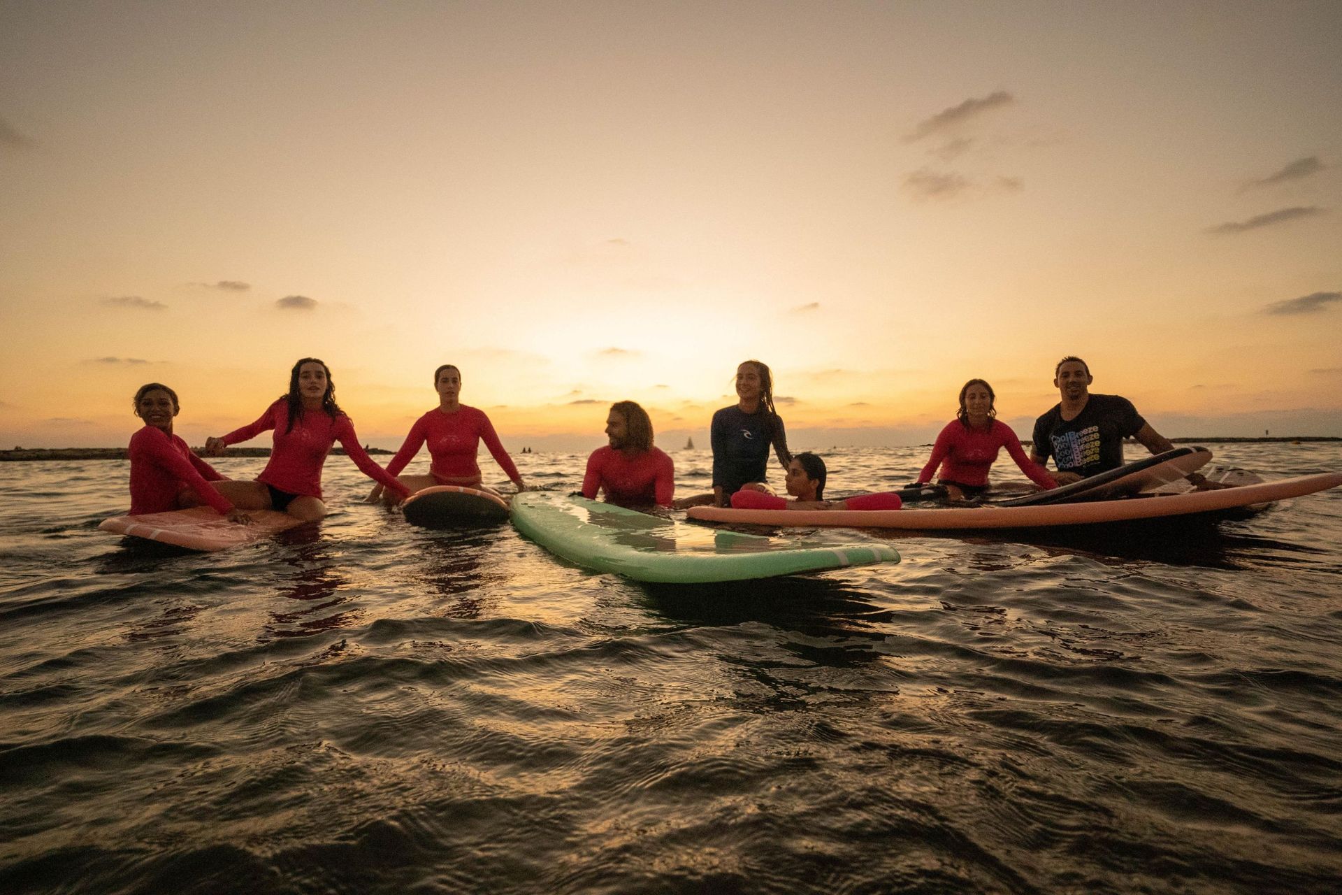 People on surfboards in the ocean at sunset; silhouetted figures, warm colors, holding hands.