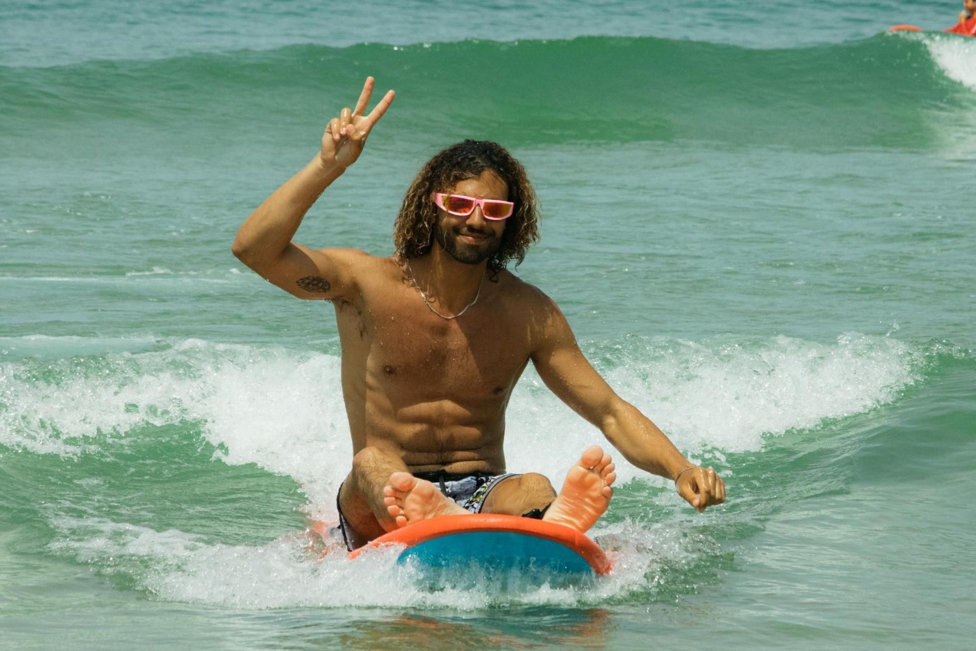Man with curly hair and sunglasses rides a skimboard in the ocean, giving a peace sign.
