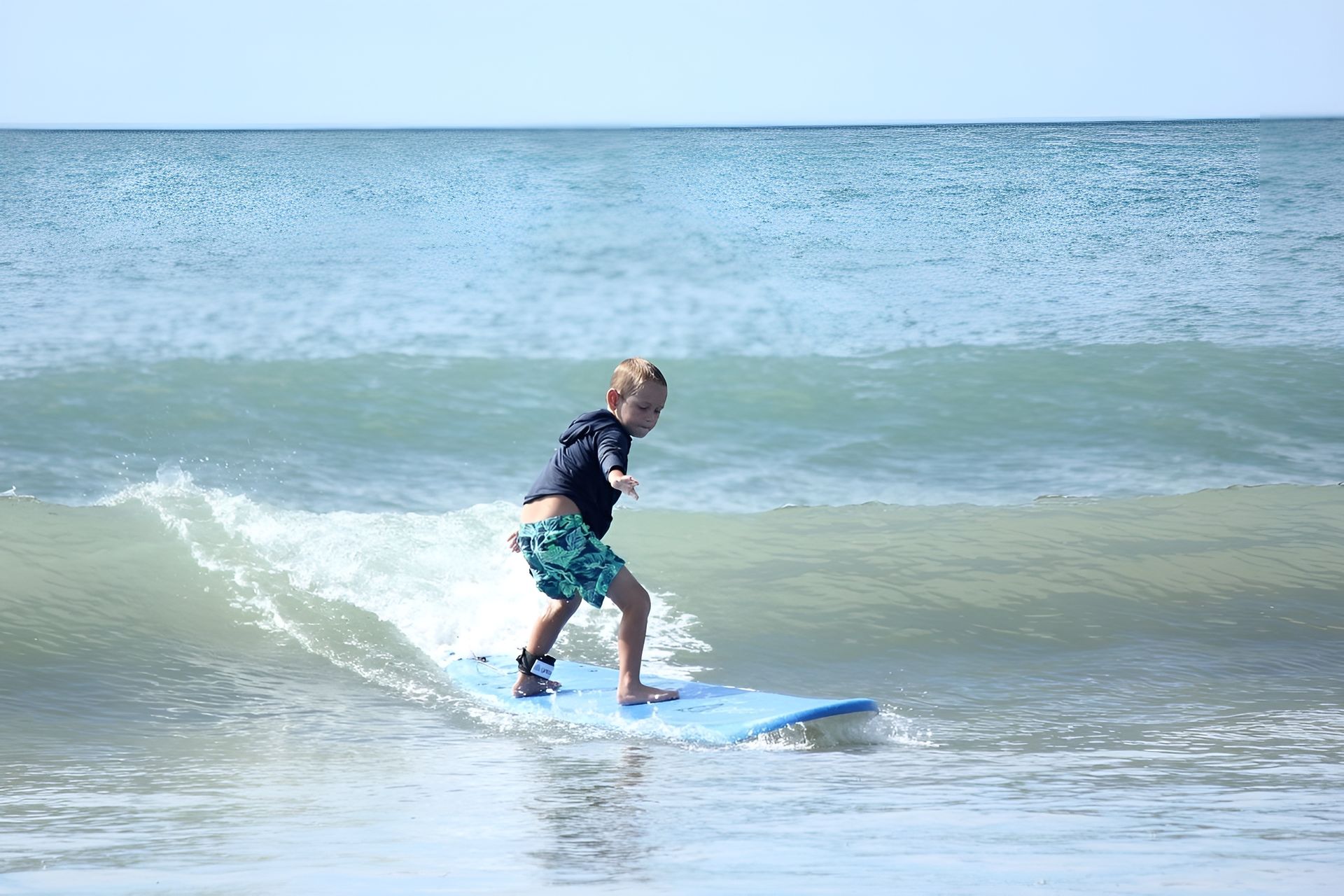 Boy surfs on a blue board in ocean, wearing swim trunks and a top on a sunny day.