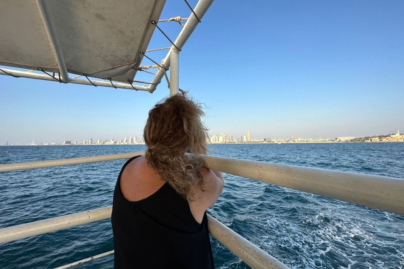 Woman on a boat looks at city skyline on a sunny day. Blue water, sky, and metal railing.