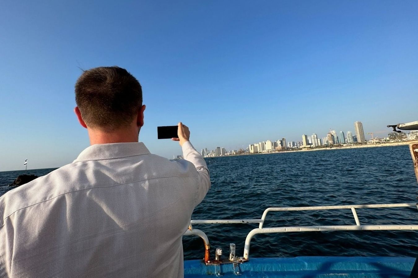 Man on a boat takes a photo of a city skyline across the blue ocean under a clear sky.
