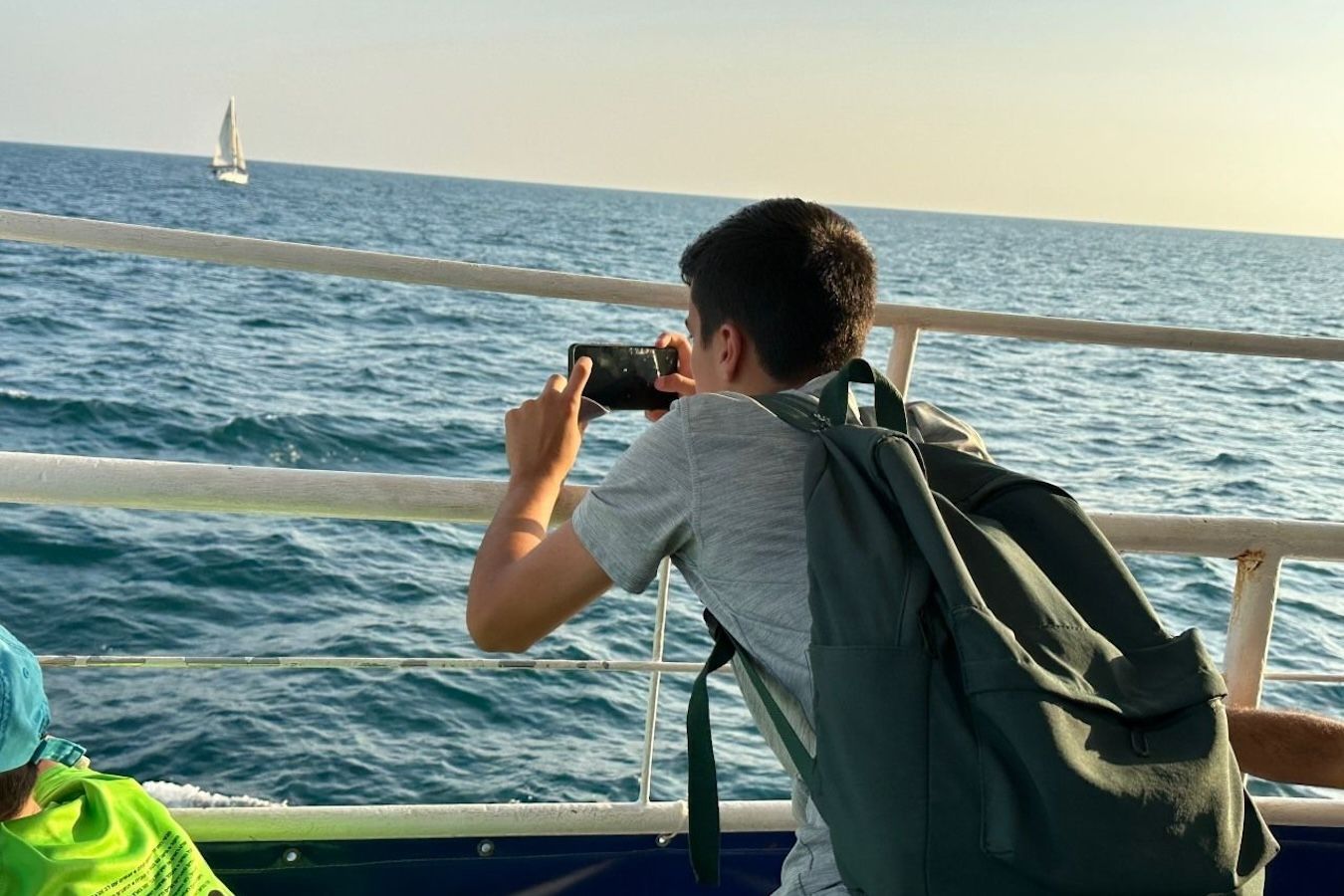 Person on a boat taking a photo of a sailboat on the blue sea, wearing a backpack.