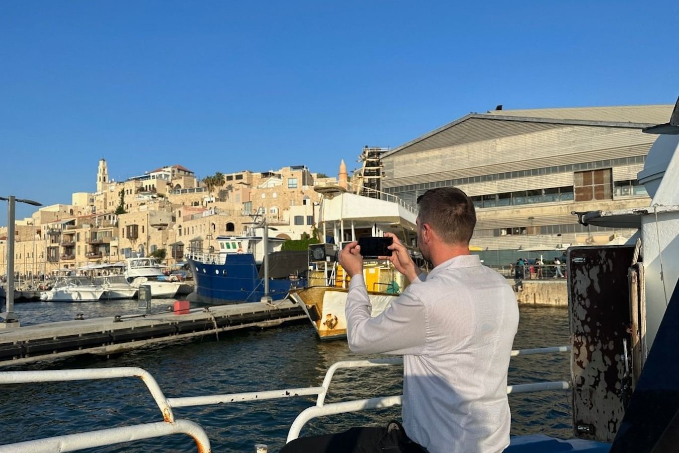 Man on a boat takes a photo of a harbor with boats, buildings, and a blue sky.