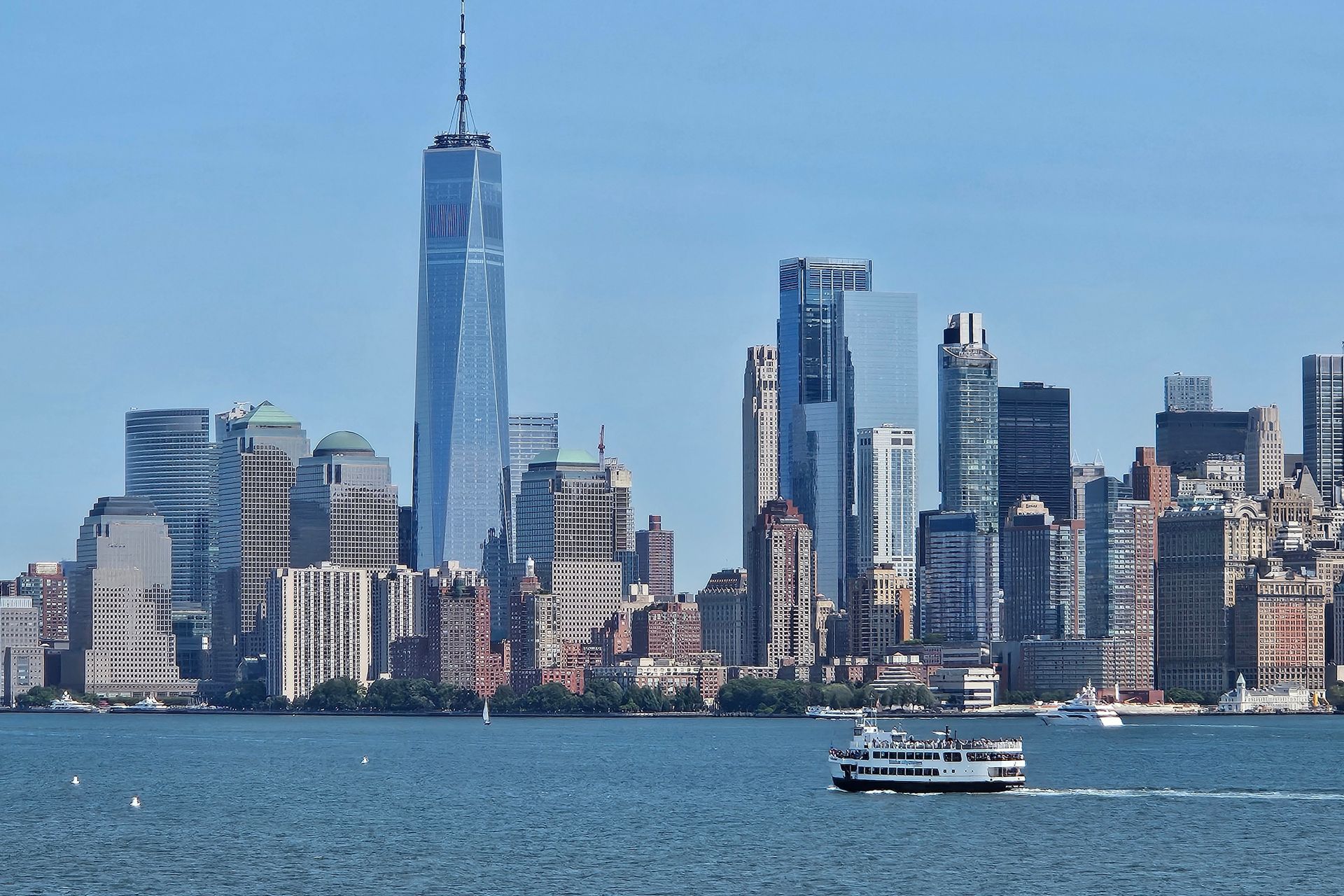 New York City skyline with the One World Trade Center, ferry on water, blue sky.