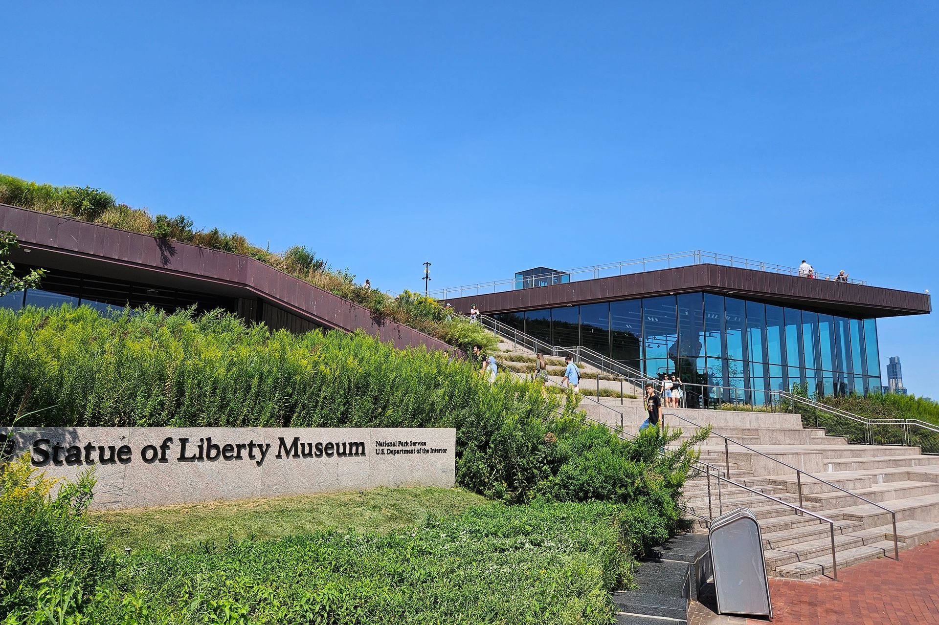 Statue of Liberty Museum entrance with sign, glass walls, and green rooftop, under a blue sky.