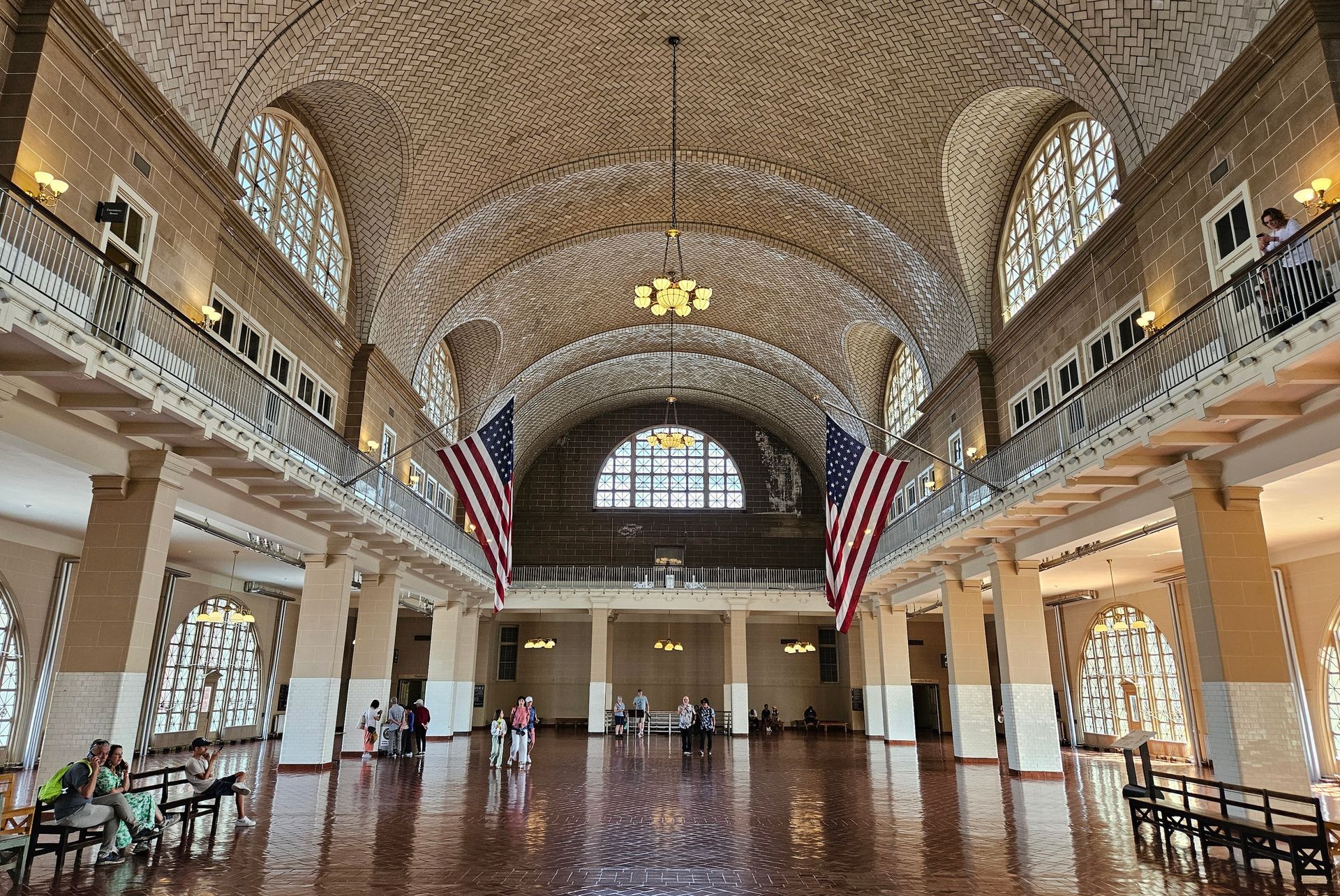 Ellis Island's Great Hall interior, brick floor, arched ceiling, American flags, windows, and people.