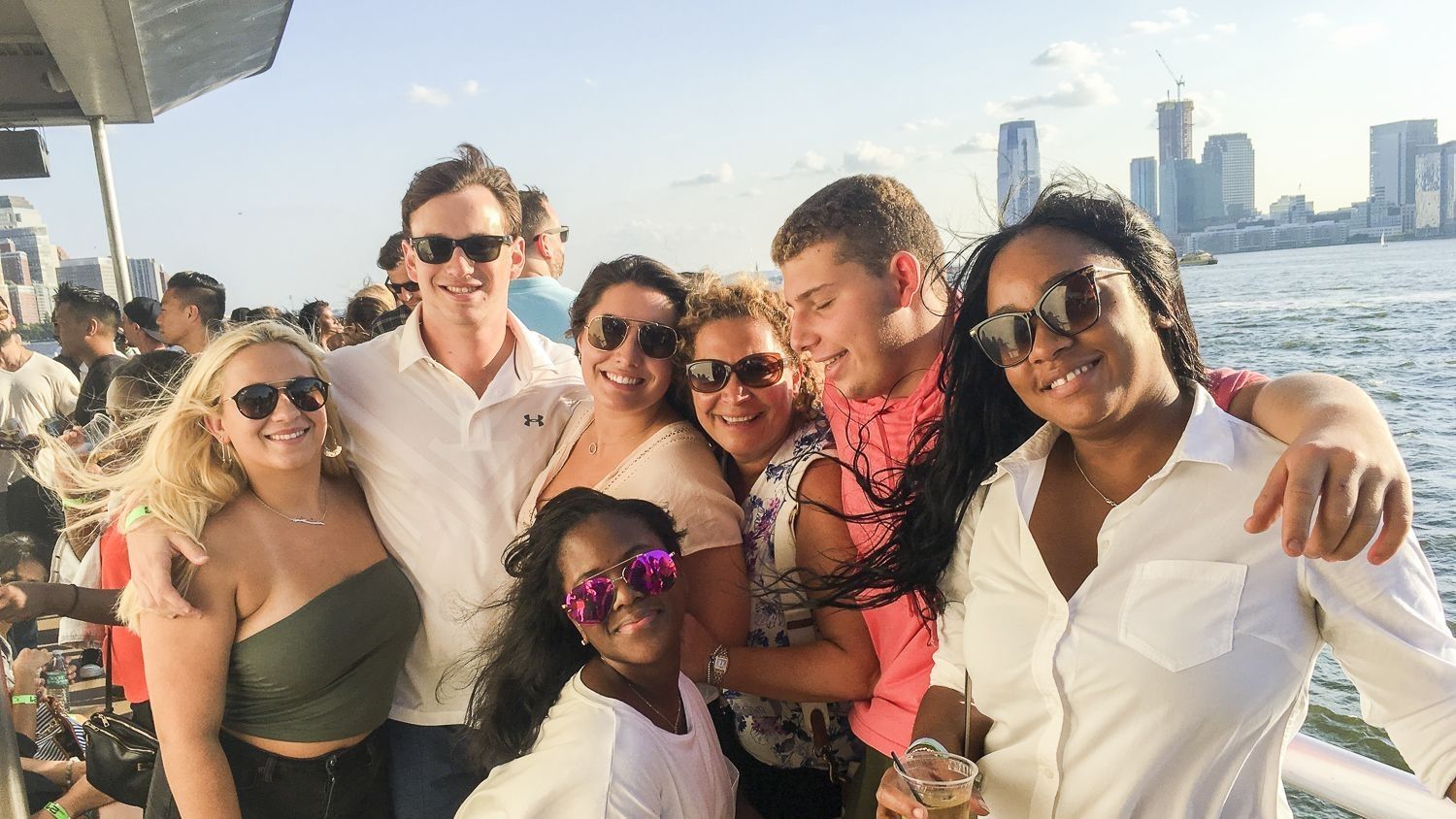 Group of people smiling on a boat, near a city skyline. Sunlight shines.