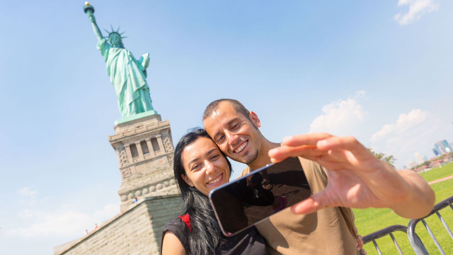 Couple taking selfie in front of the Statue of Liberty, blue sky background.