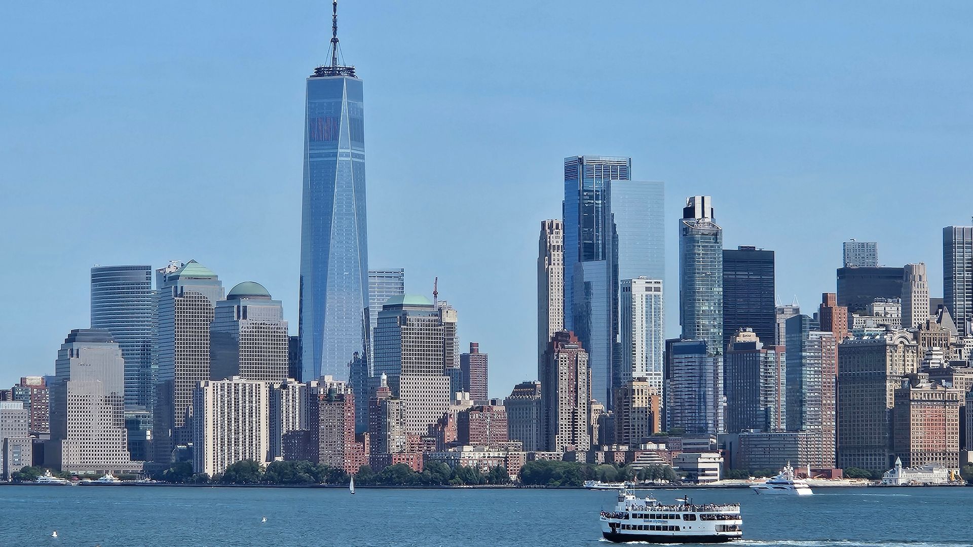 New York City skyline with the One World Trade Center tower and a ferry on water under a blue sky.