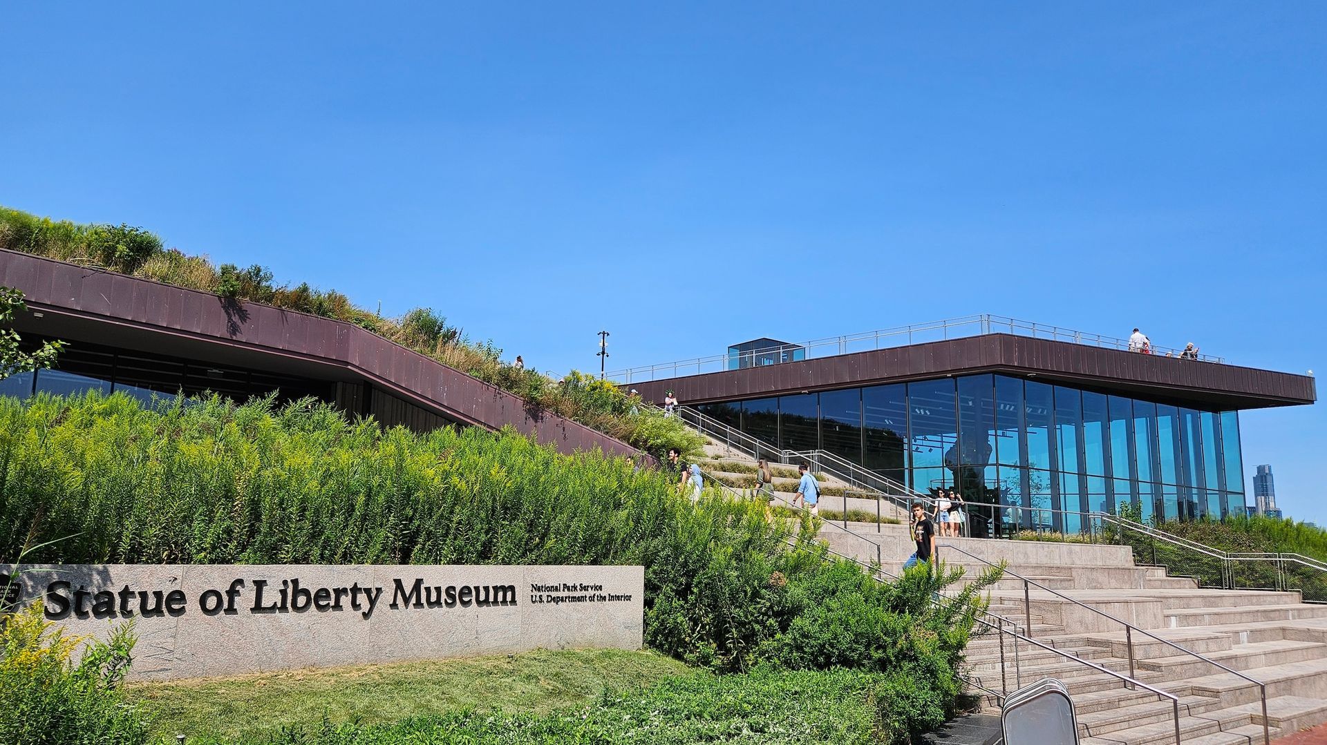 Statue of Liberty Museum exterior with glass facade, green bushes, and blue sky.