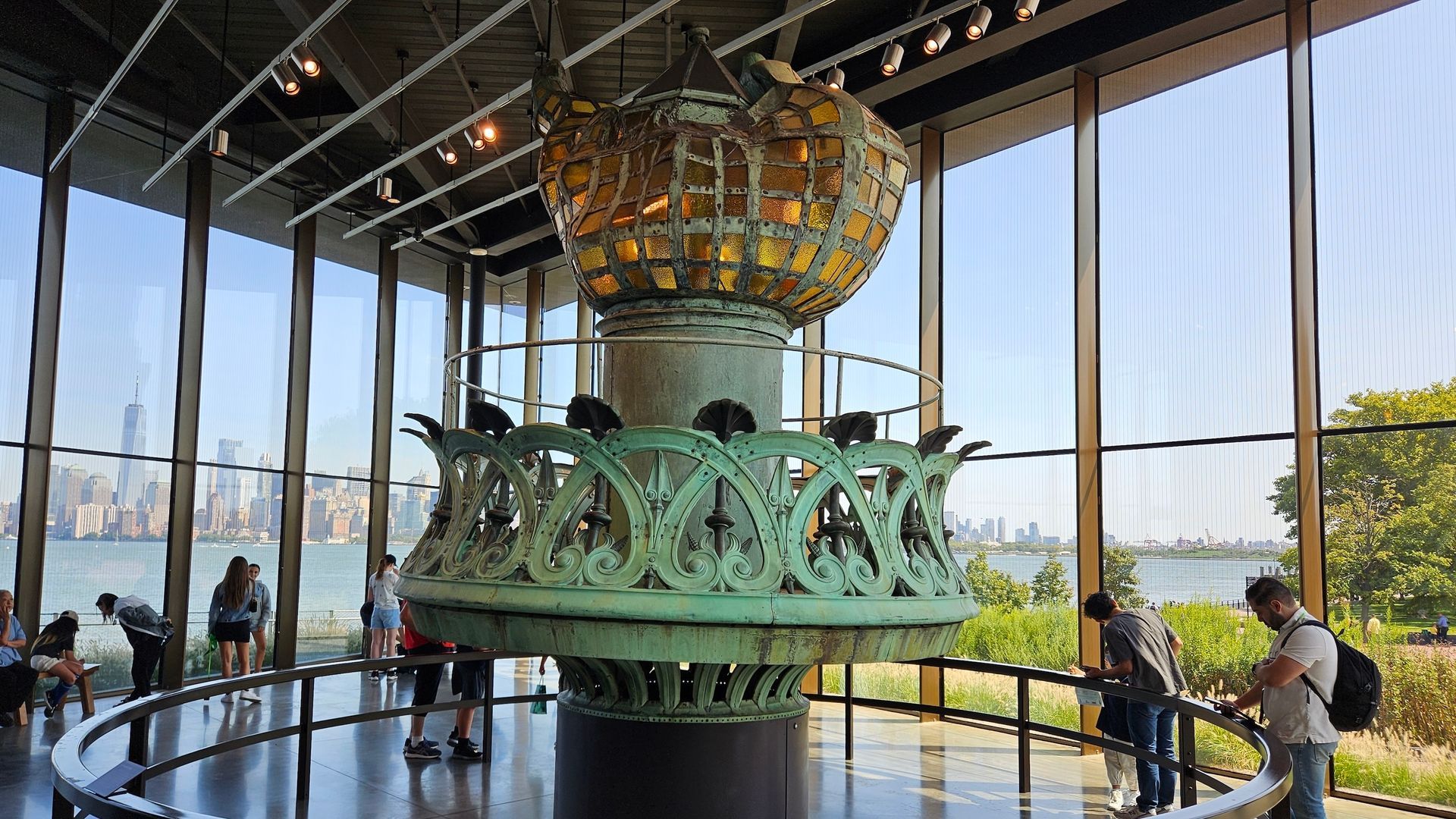 Statue of Liberty's original torch on display in a museum, people viewing; New York City harbor visible.