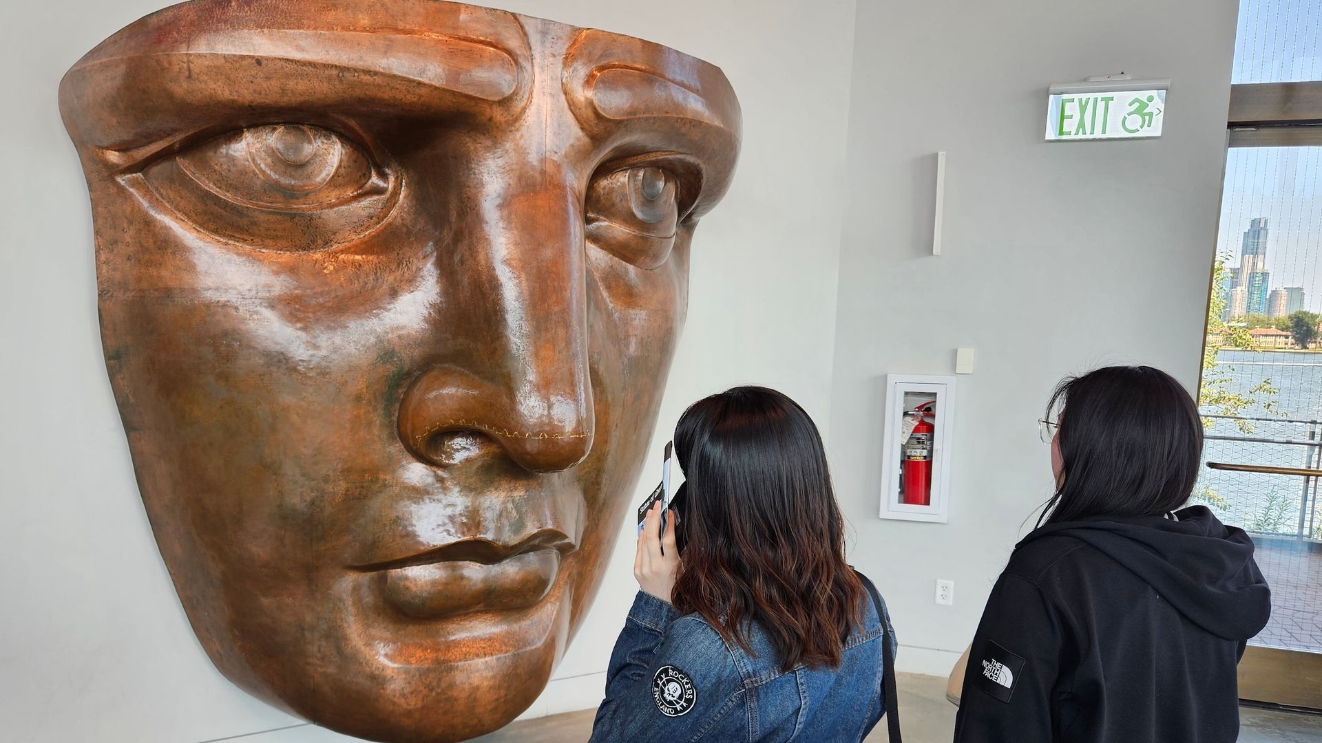Two women looking at a large, copper-colored sculpture of a face on a white wall, inside a building.