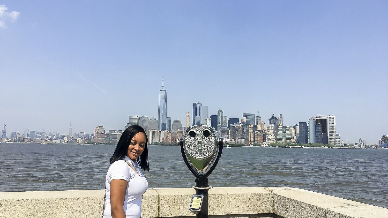Woman poses in front of NYC skyline near a viewing telescope.