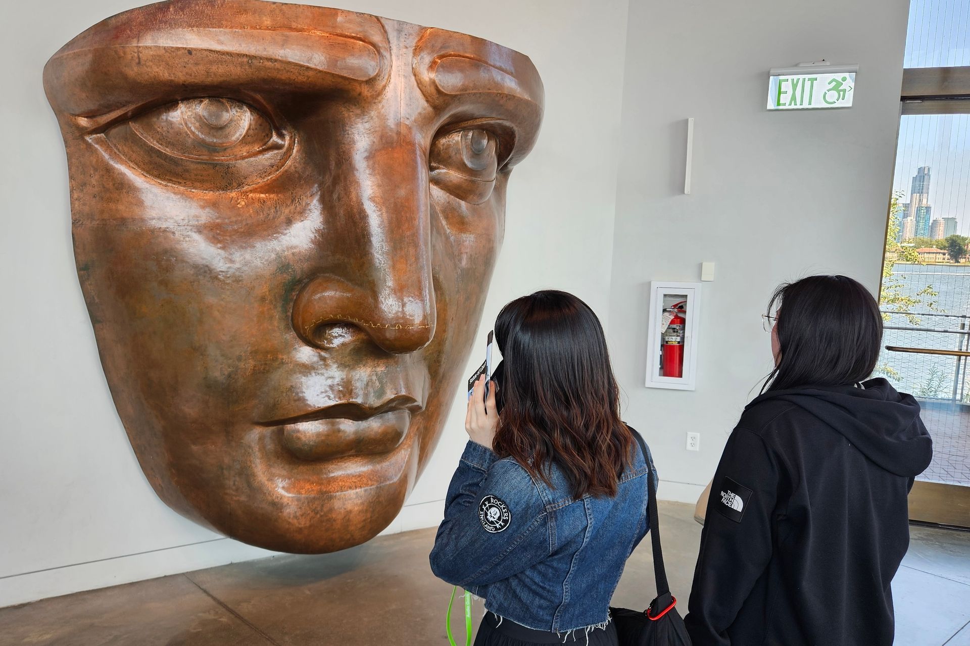 Two women looking at a large, bronze sculpture of a face mounted on a wall inside a museum.