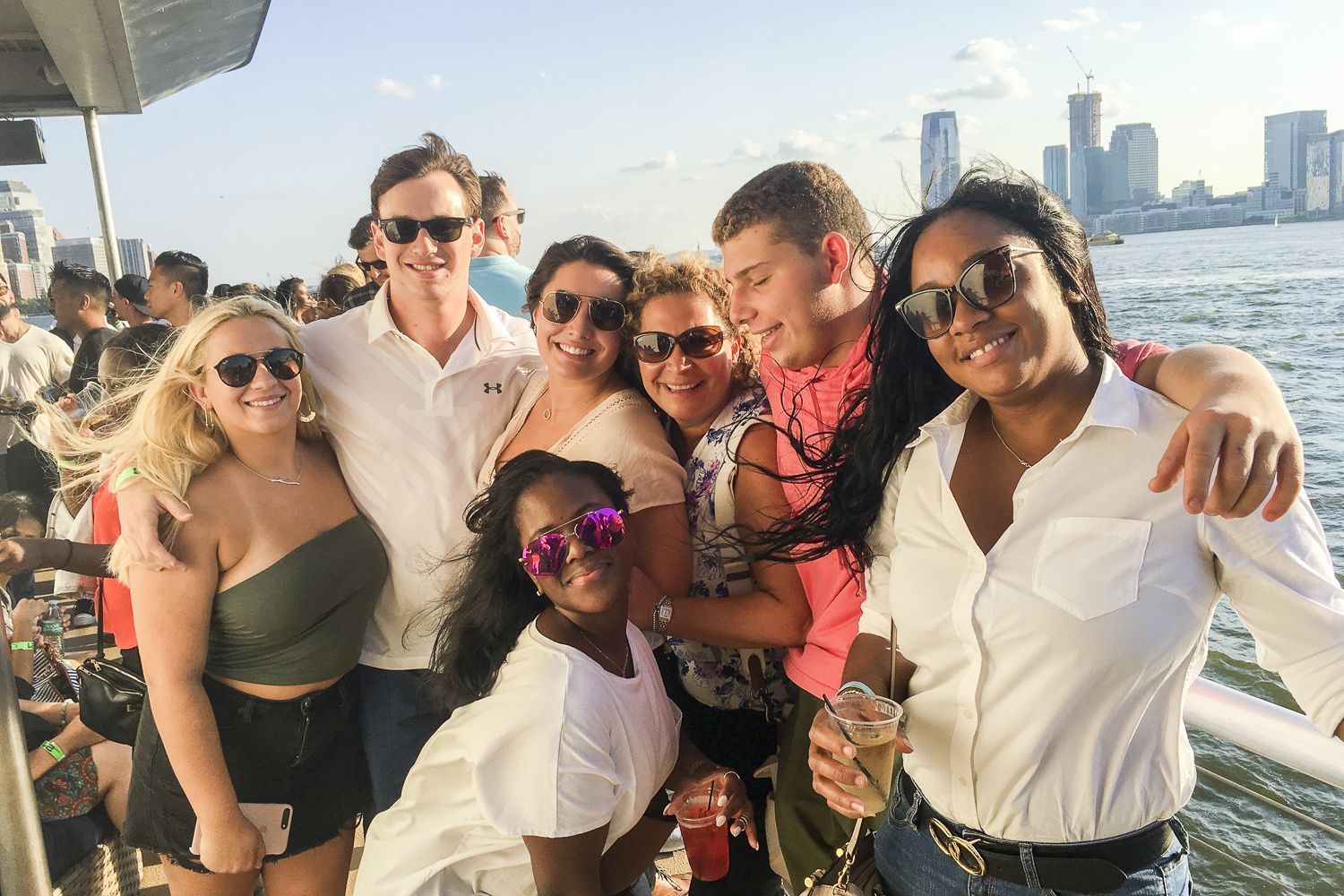 Group of diverse friends smile and pose on a boat; city skyline in the background.