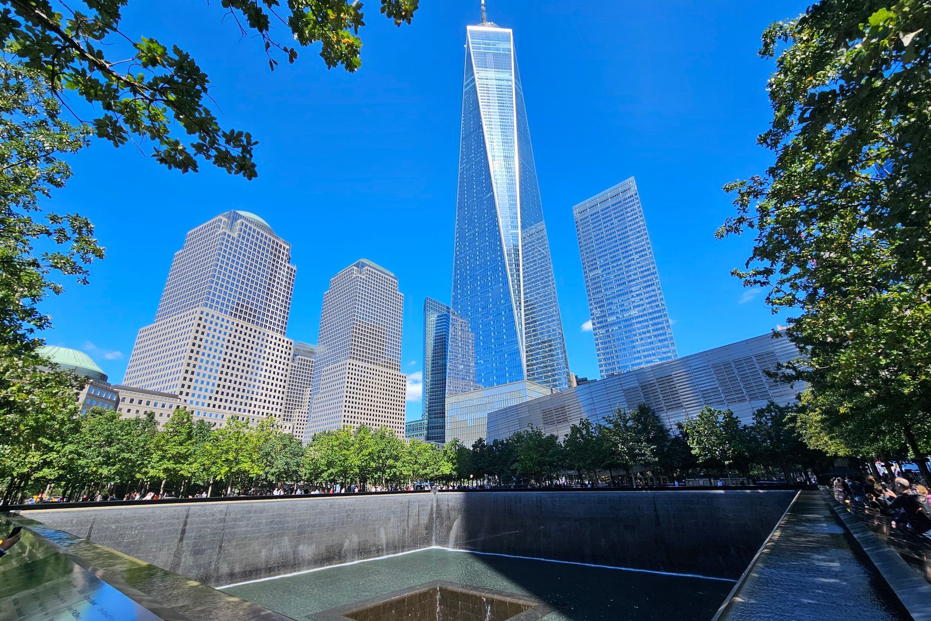 One World Trade Center and other skyscrapers rise above the 9/11 Memorial pools, framed by trees, against a clear blue sky.