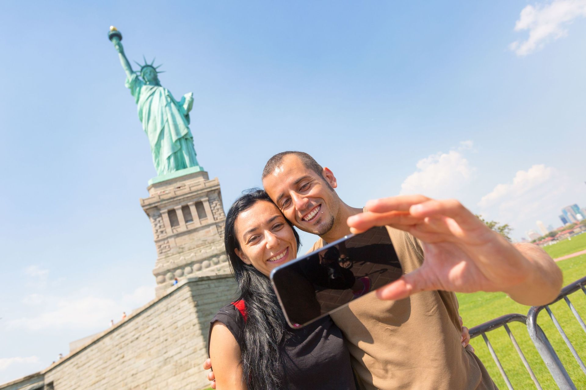 Couple taking selfie with Statue of Liberty in background, sunny day.