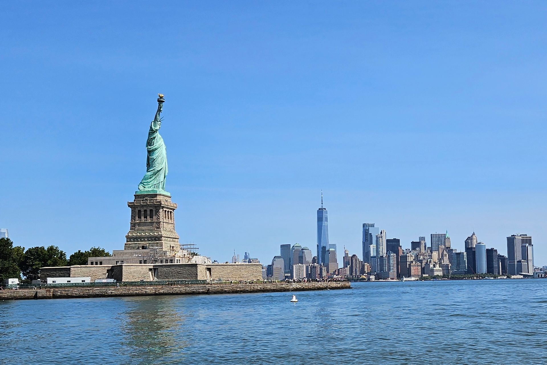 Statue of Liberty stands on an island, facing the NYC skyline across the blue water on a sunny day.