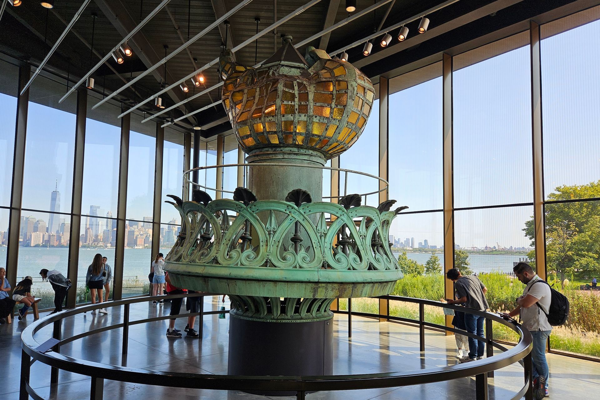 The Statue of Liberty's torch in a museum; people view it near a window with a city skyline.