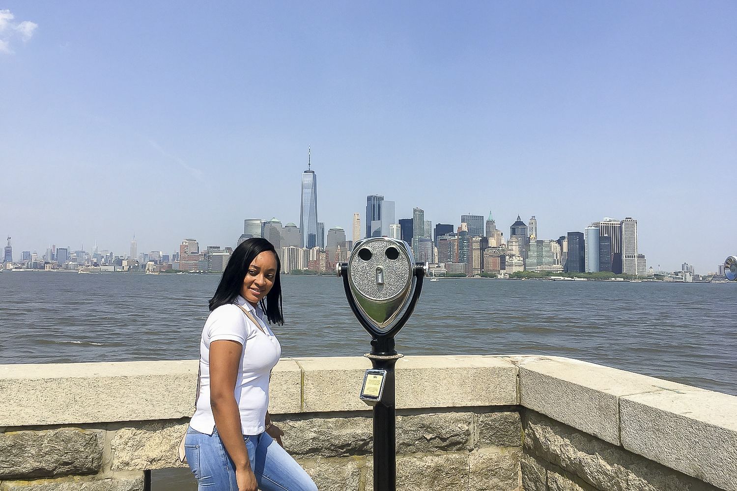 Woman smiles at camera in front of the NYC skyline and a coin-operated viewer on a sunny day.