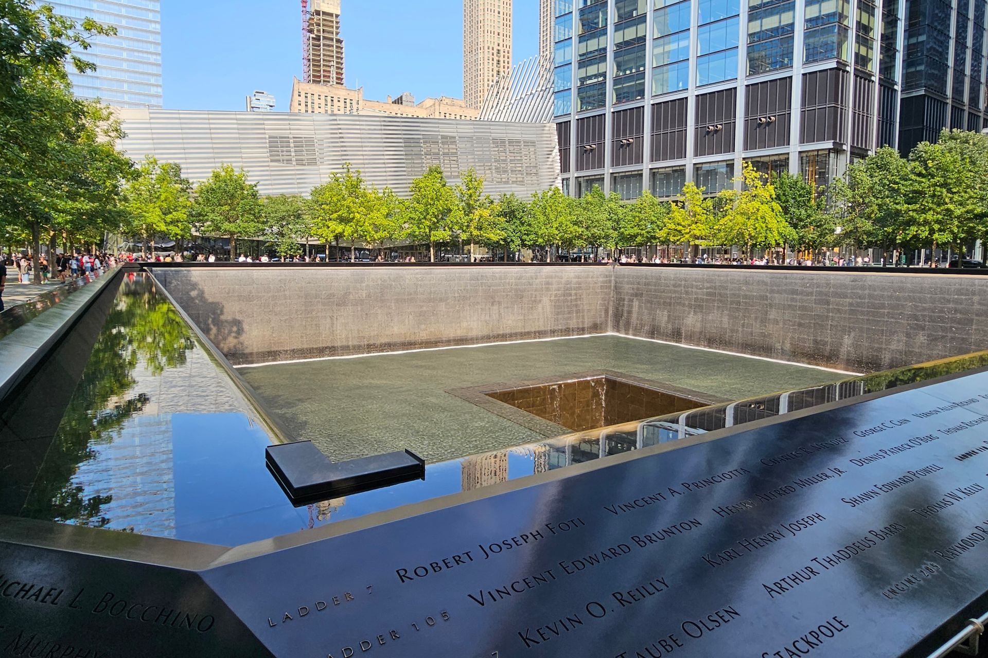 9/11 Memorial reflecting pool with names etched, skyscrapers, and green trees in New York City.