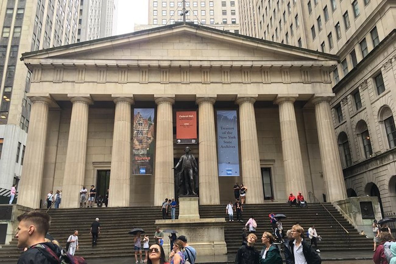 Building with columns and statue, people walking and sitting outside in New York City.