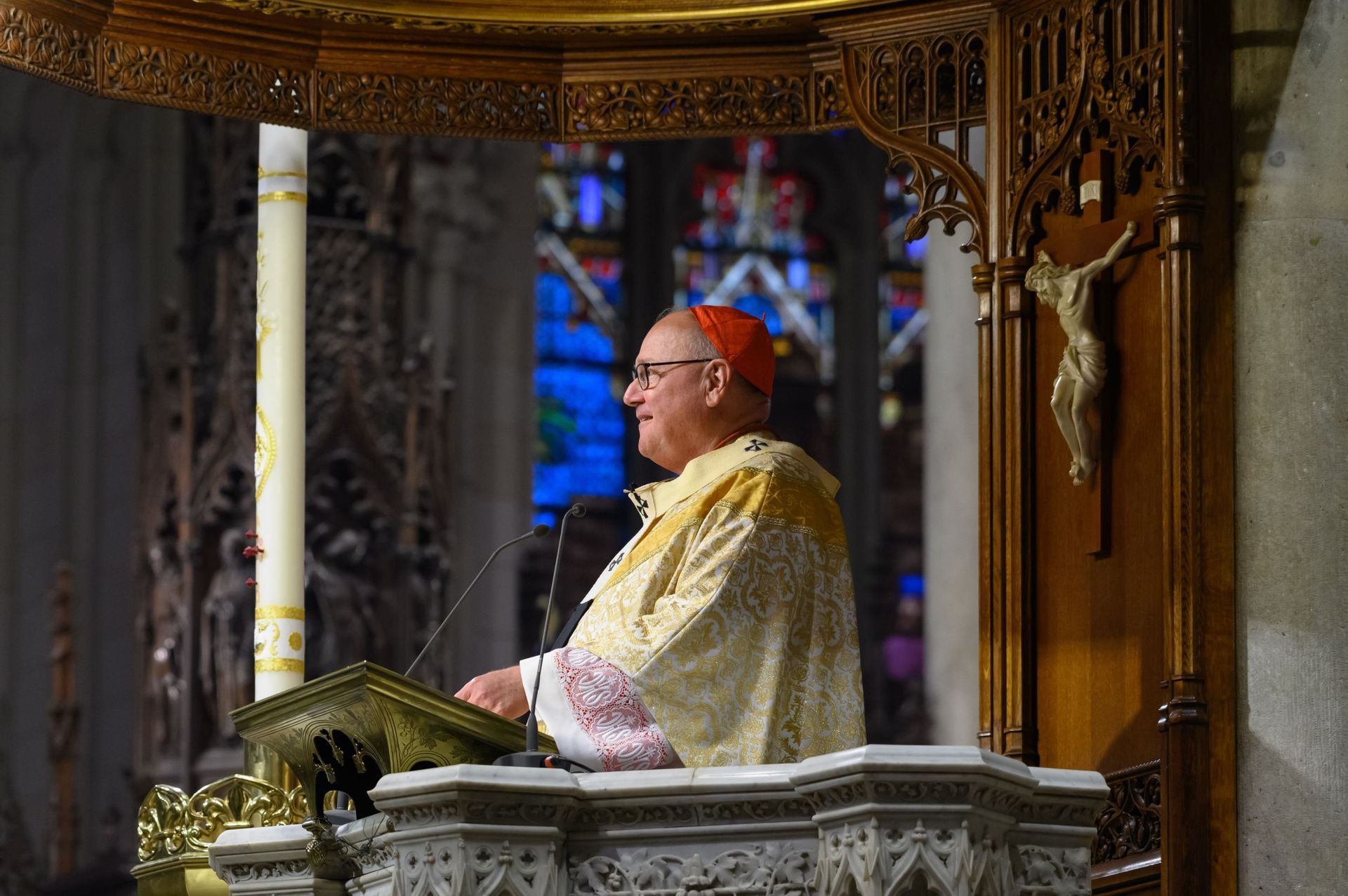 Cardinal speaking from a pulpit inside a church, wearing red and gold vestments.