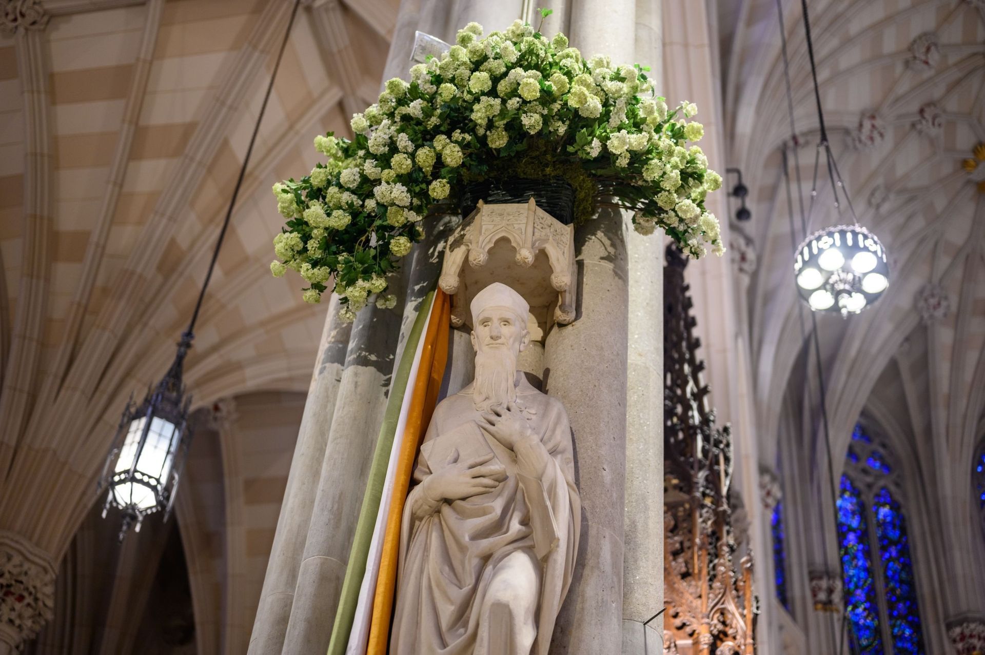 Statue of a saint in a cathedral, surrounded by flowers. Pale stone, white roses, arched architecture.