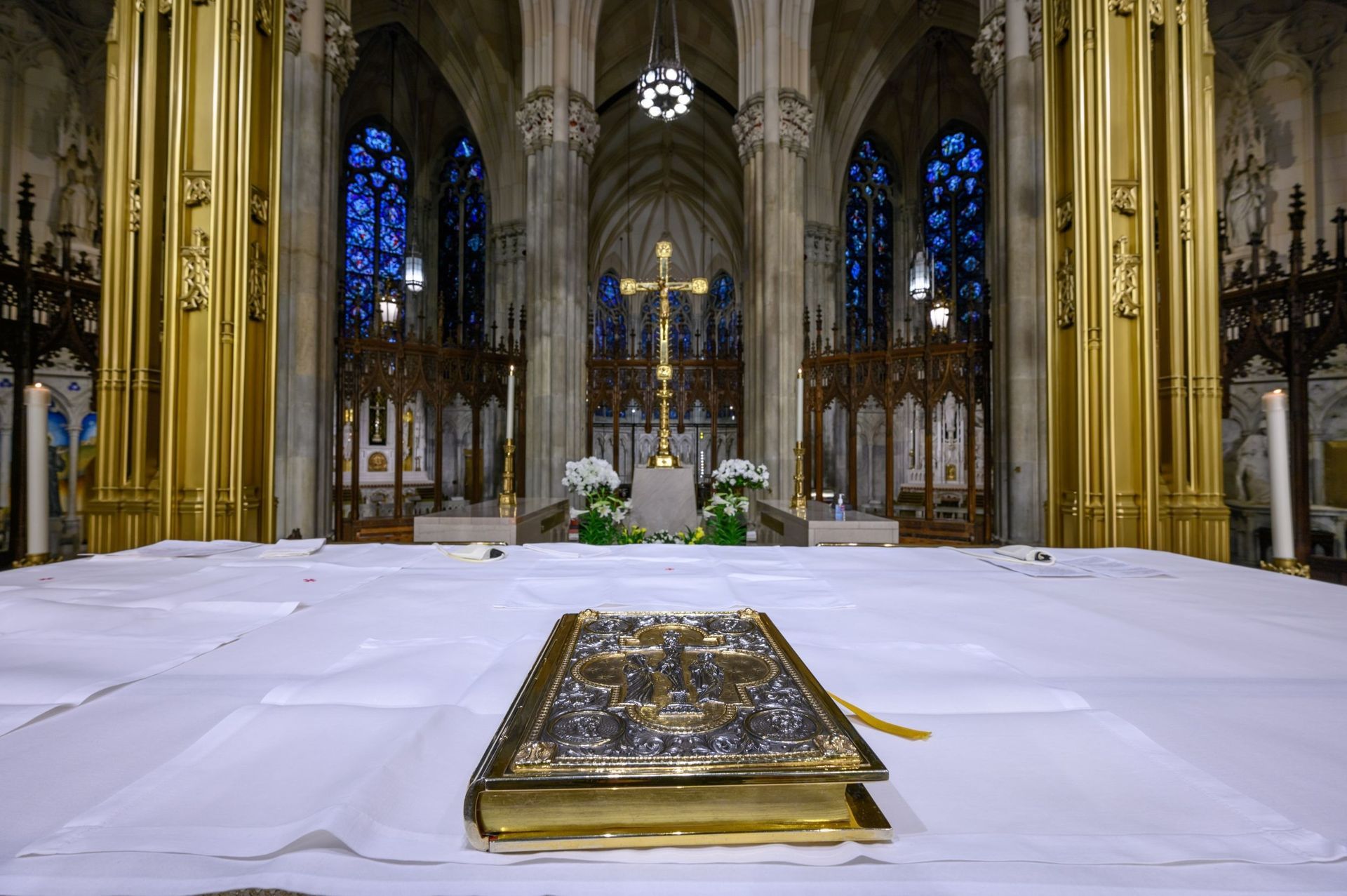 Inside a cathedral, a gold book on a white tablecloth. A gold cross and altar are visible.