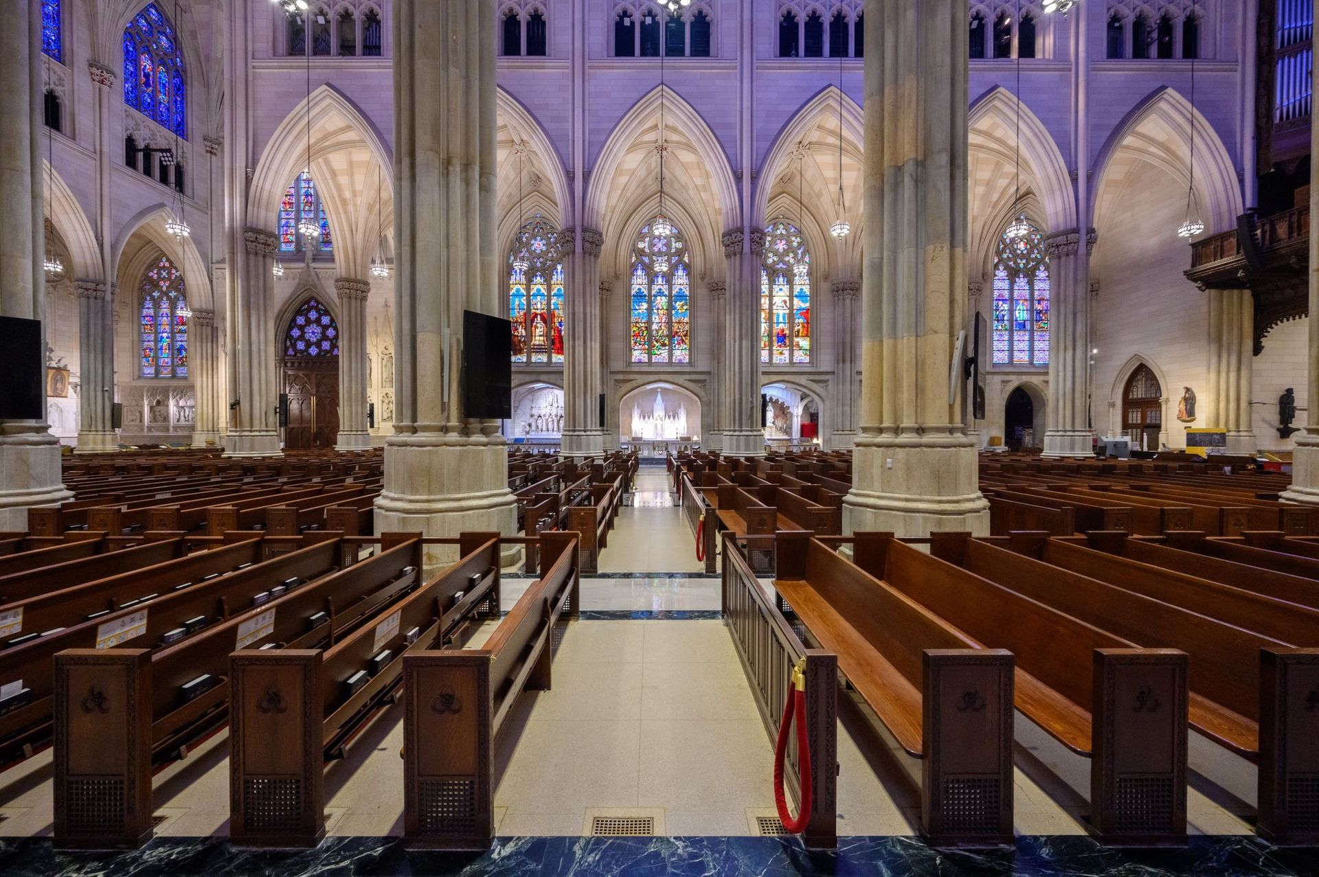 Interior of a cathedral with rows of wooden pews, stained glass windows, and tall columns.