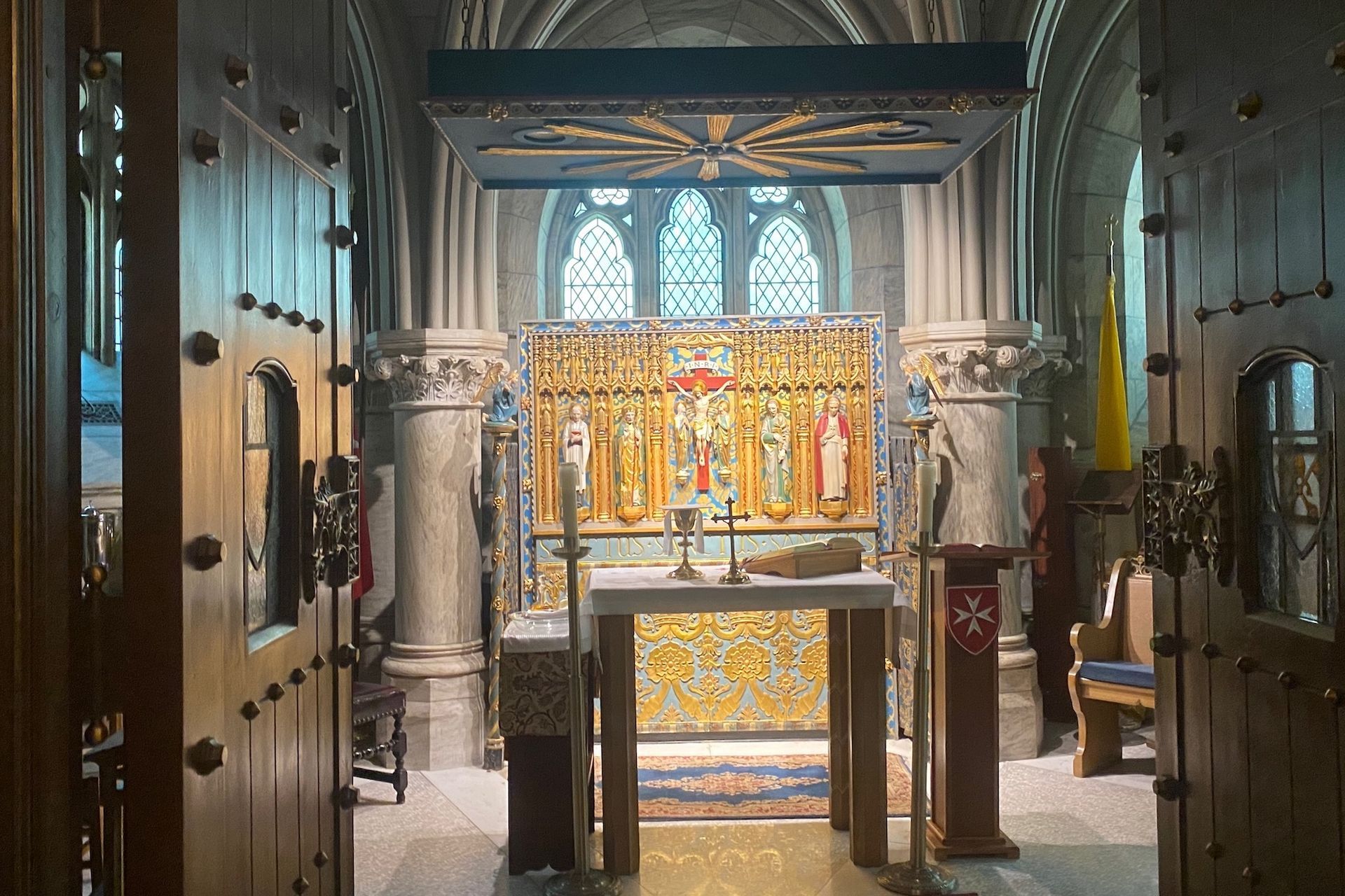 Church altar: ornate gold and stone decorations, stained glass, open wooden doors, and warm lighting.