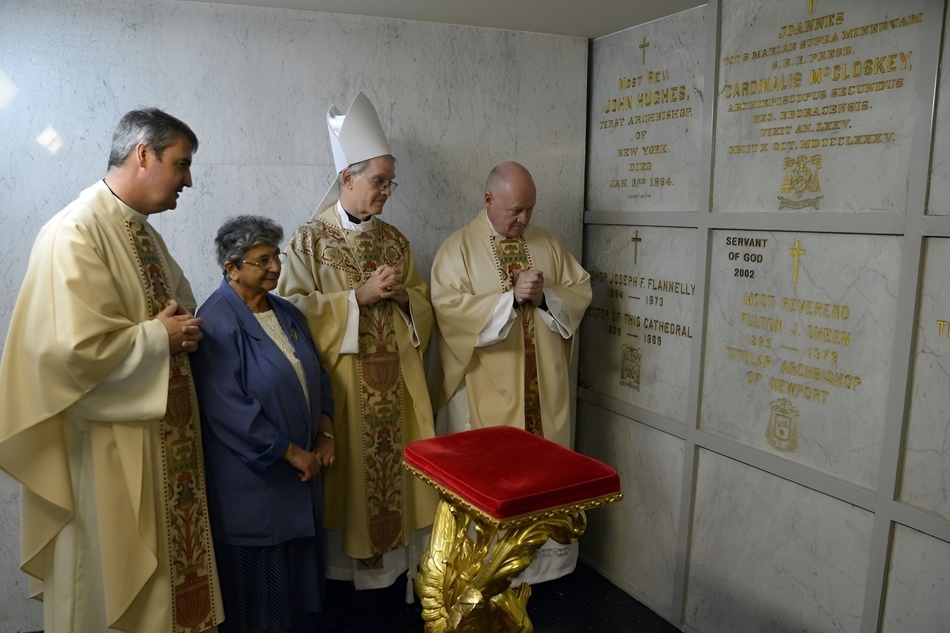 Clergy and woman praying in a mausoleum. White walls, marble crypts, golden vestments, and a red cushion.