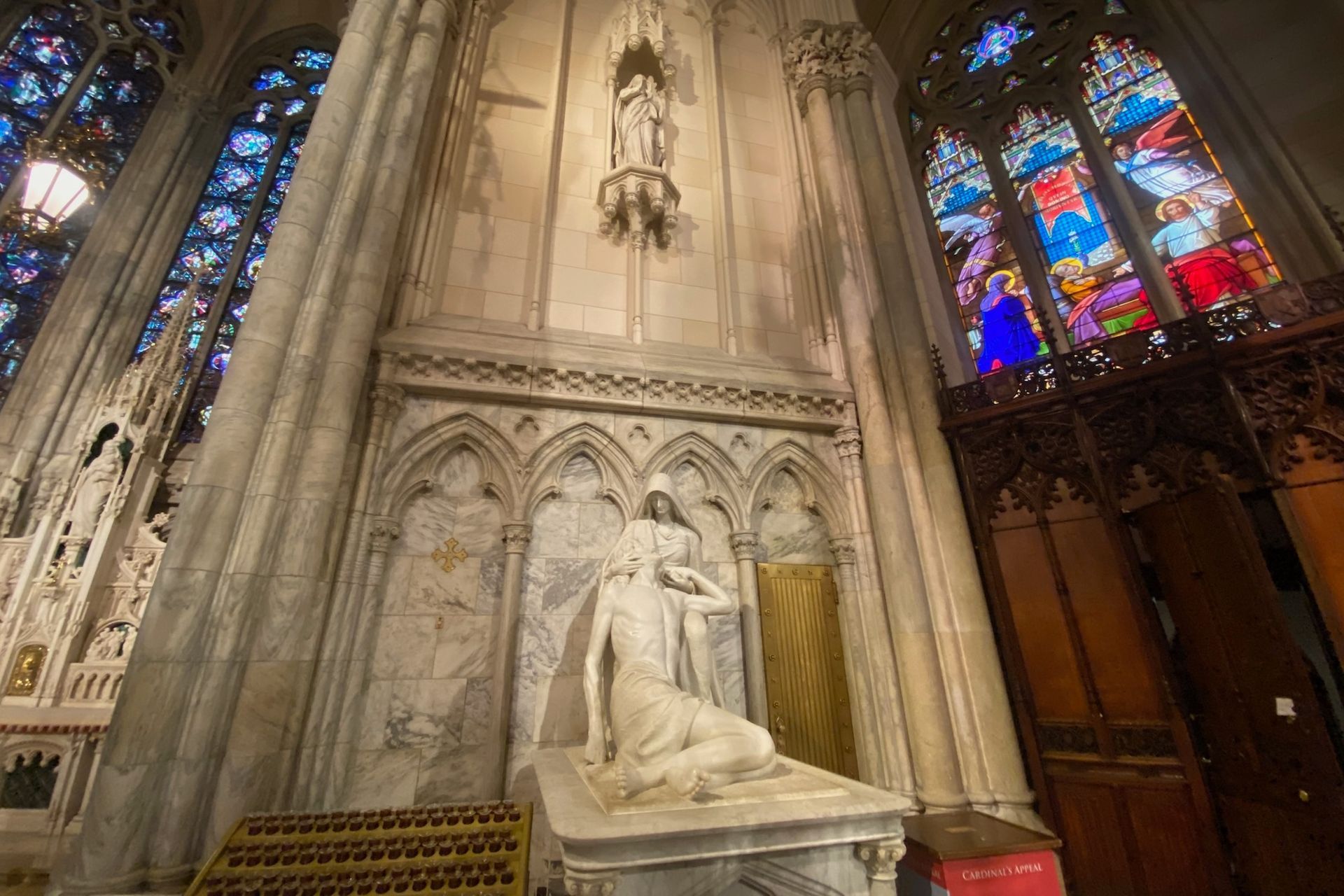 Sculpture of a figure kneeling before a standing person in a church with stained glass windows.