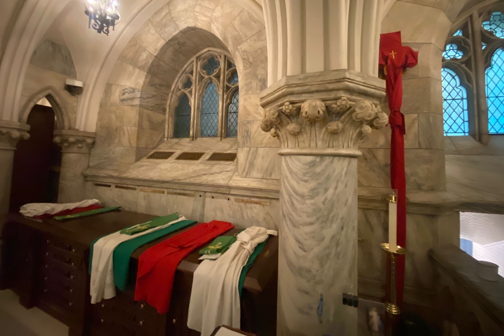 Interior of a church crypt with a marble pillar, altar, and window with stained glass. Flags of green, red and white.