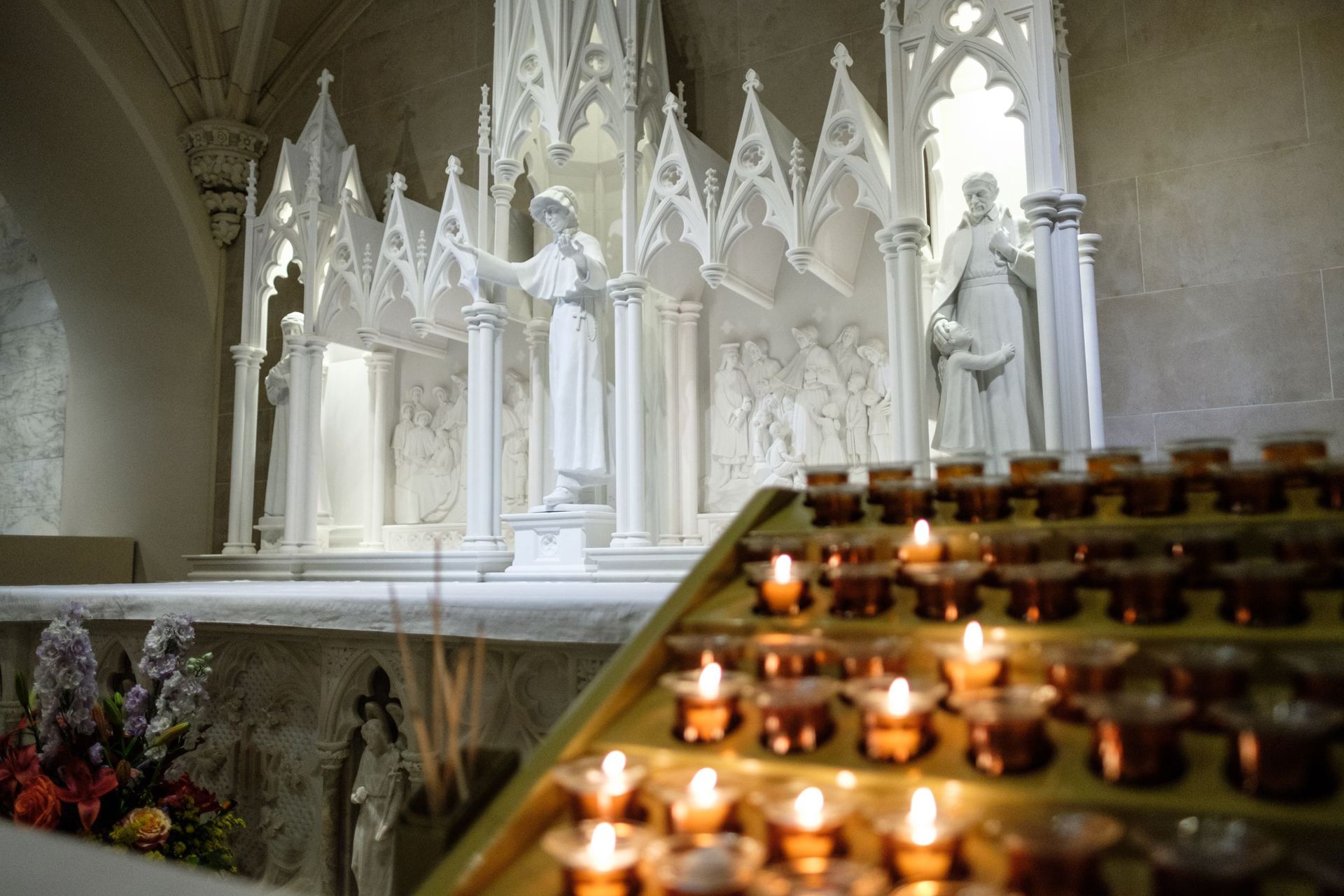 Altar with white statues, candle stand with lit candles, and floral arrangement in a church.