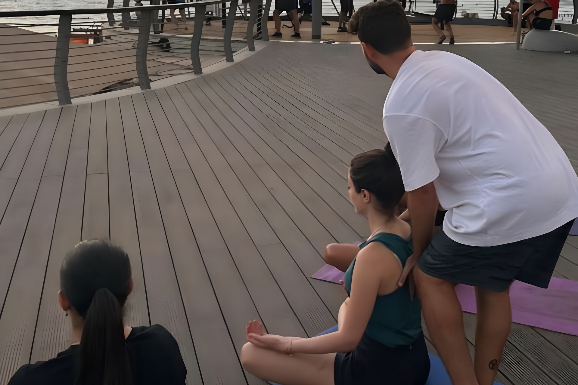 Yoga instructor assisting a student outdoors. Instructor in white shirt, student in teal top. On wooden deck.